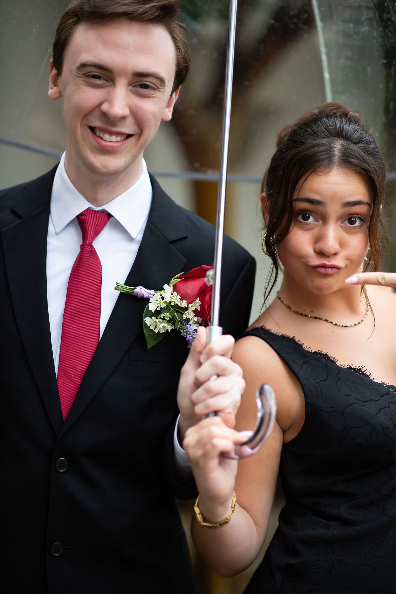 Smiling groom in black suit with red tie and boutonniere standing with bridesmaid under clear umbrella.