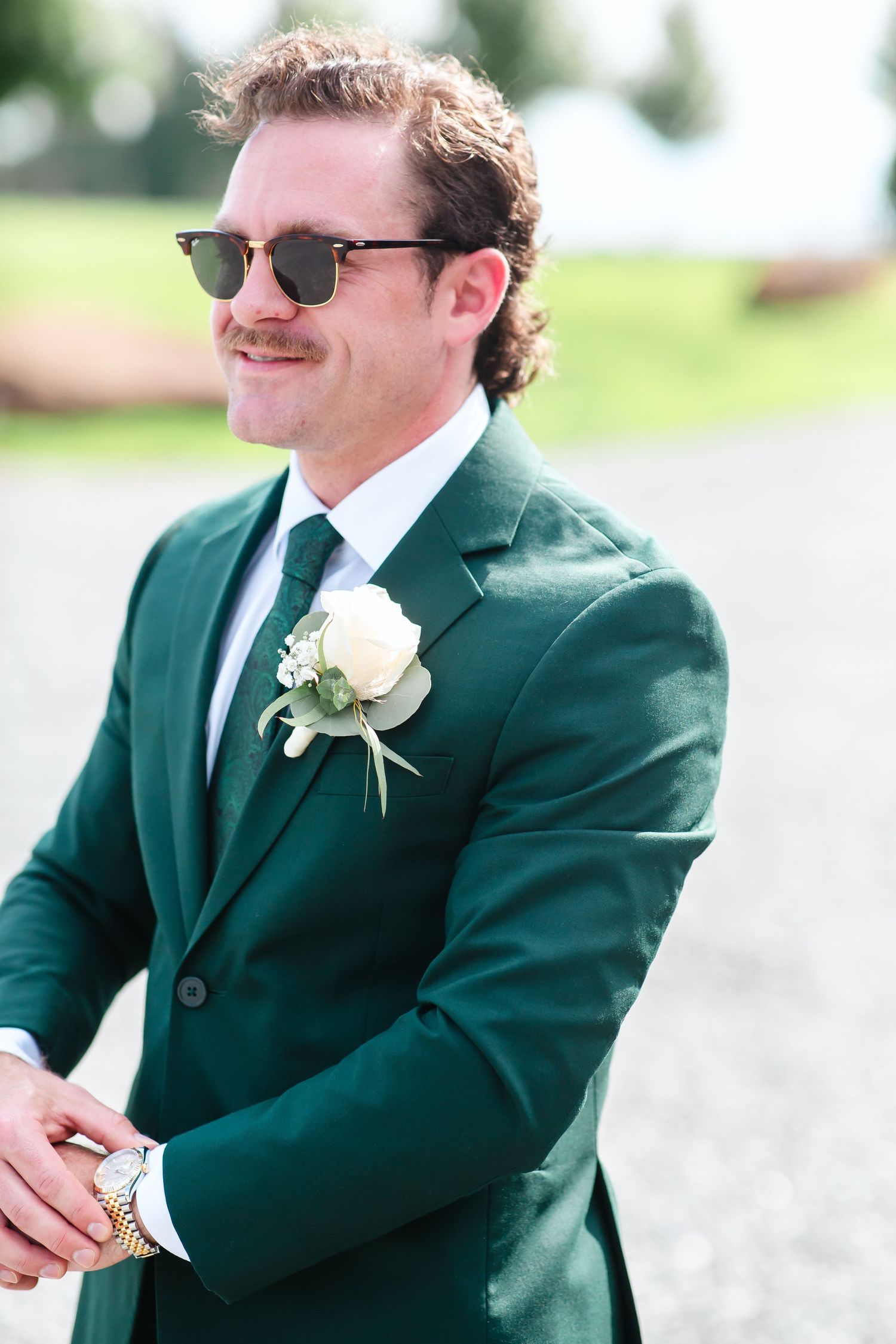 Groom in emerald green suit with white boutonniere and sunglasses smiling outdoors on wedding day.