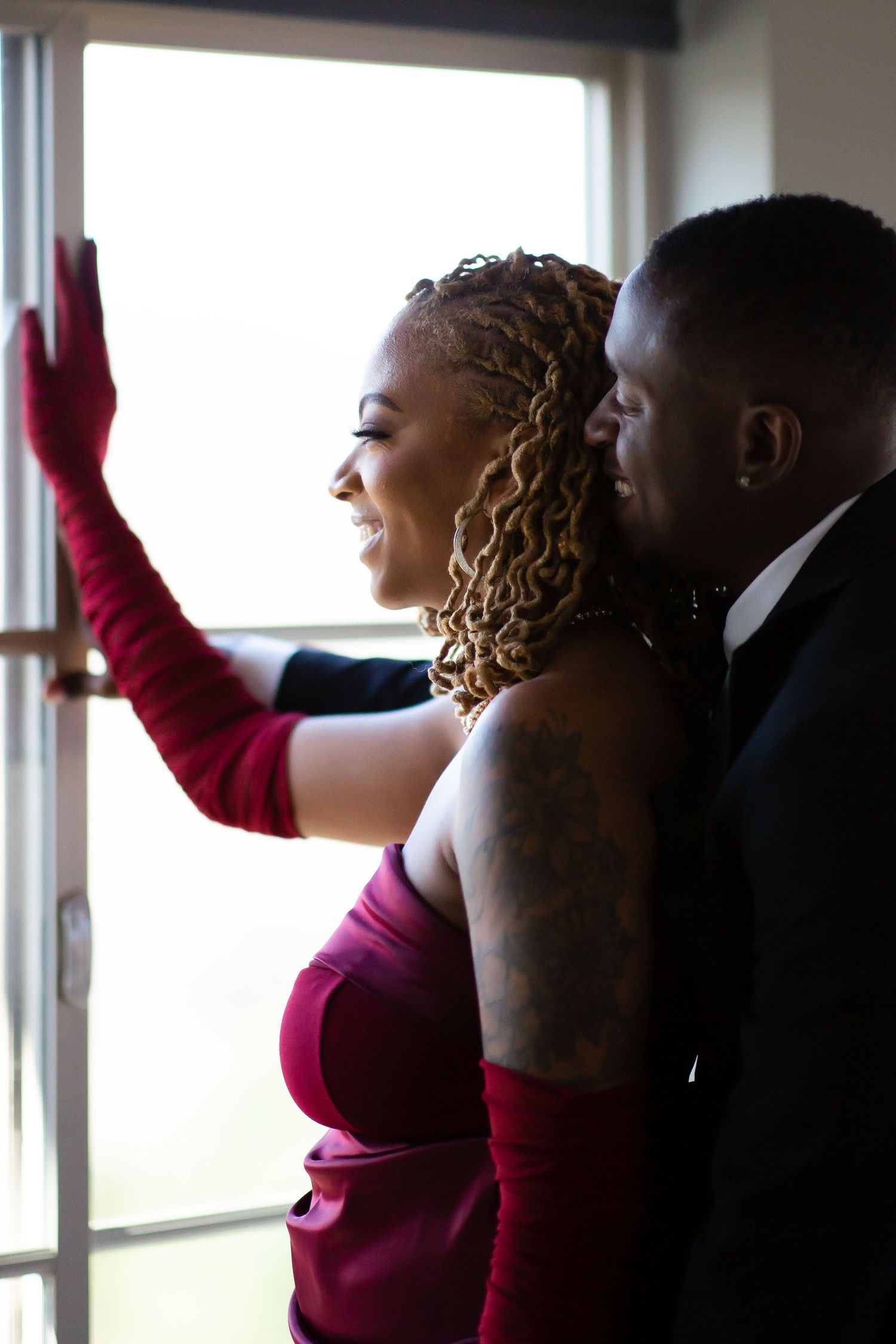 Elegant couple in formal wear sharing a joyful moment by a bright window, woman in burgundy dress smiling.