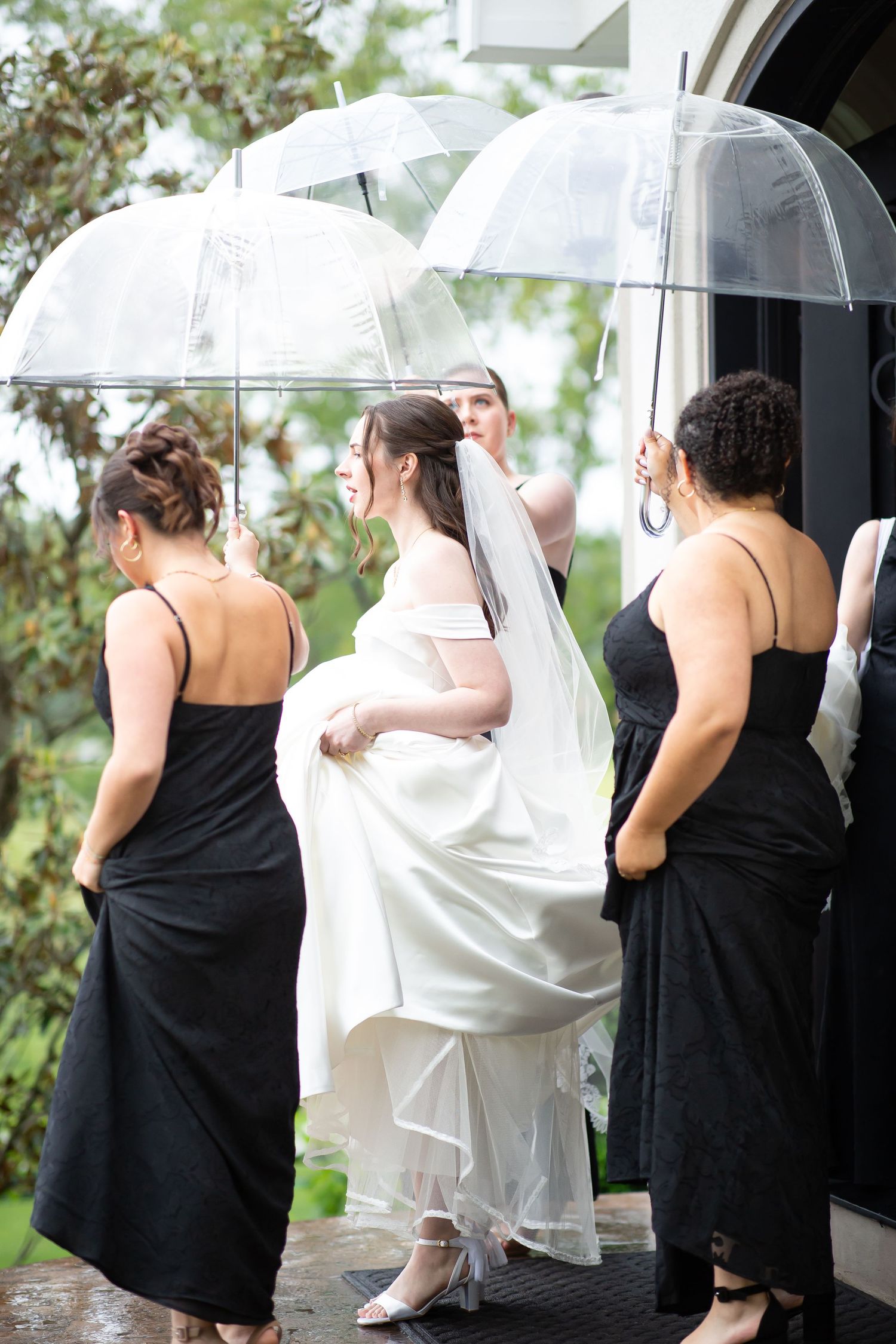 Bride in white gown with bridesmaids in black dresses holding clear umbrellas entering church in rain.