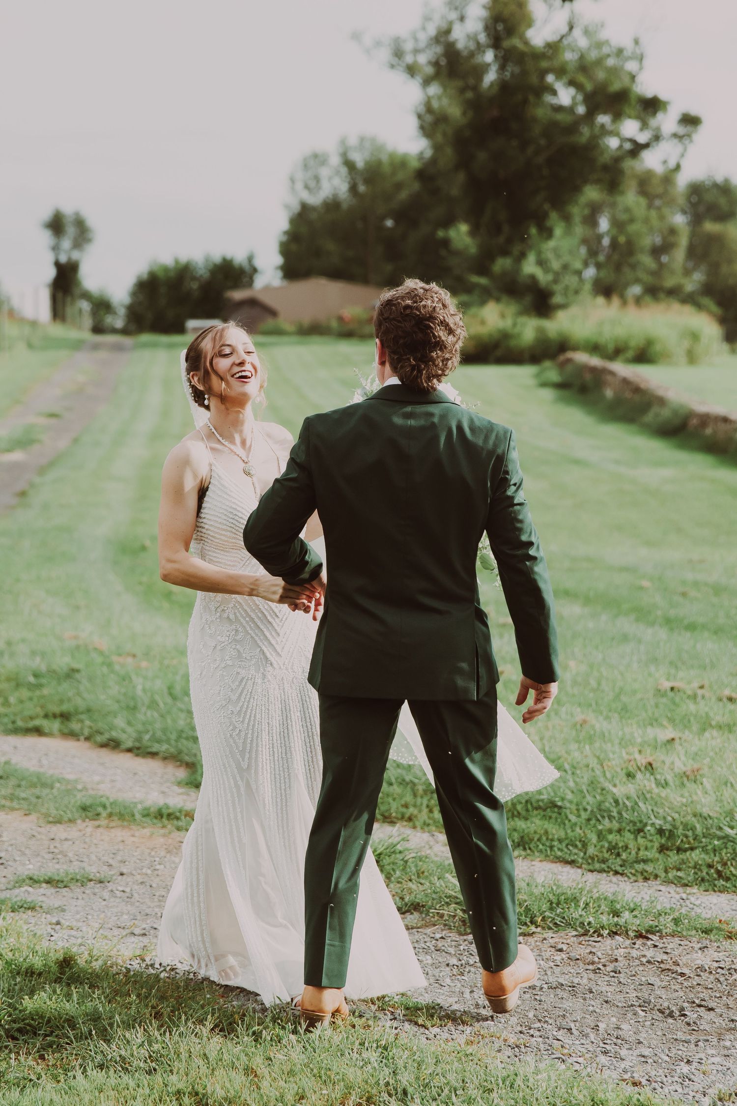 Bride in white gown laughs joyfully while walking with groom in dark suit on a grassy path.