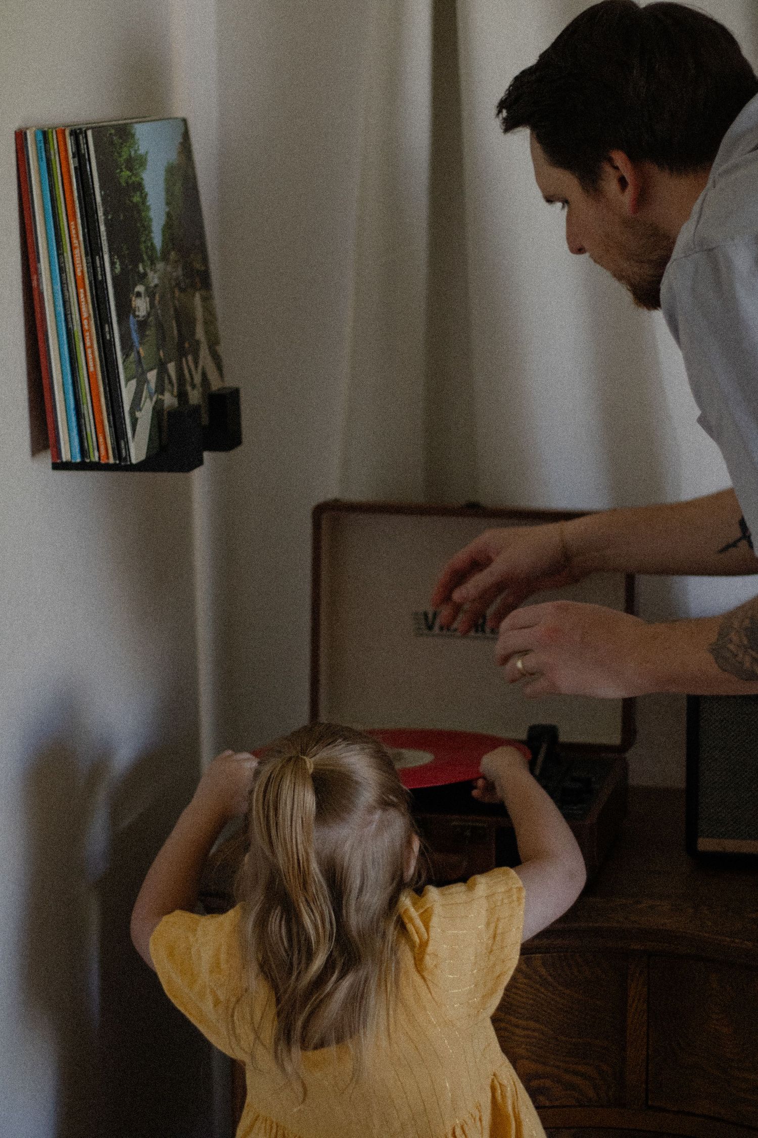 A father and young girl in yellow dress explore vinyl records on a turntable at home.