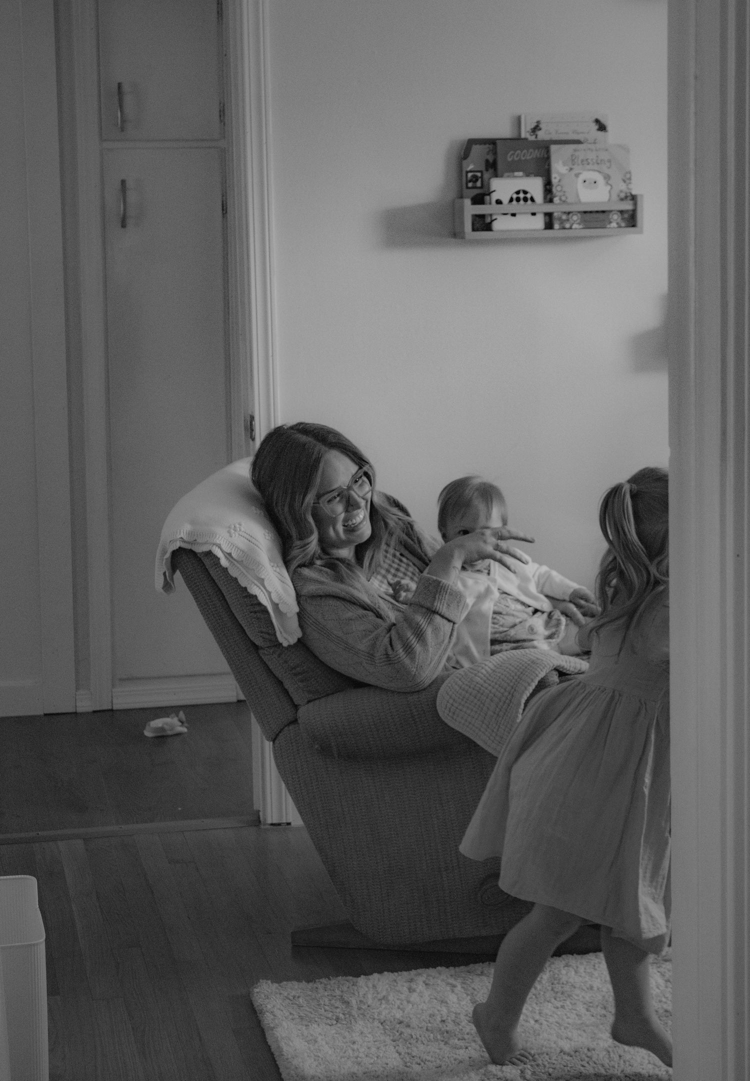 Black and white photo of a smiling mother sitting in a chair holding a baby while a young child stands nearby.