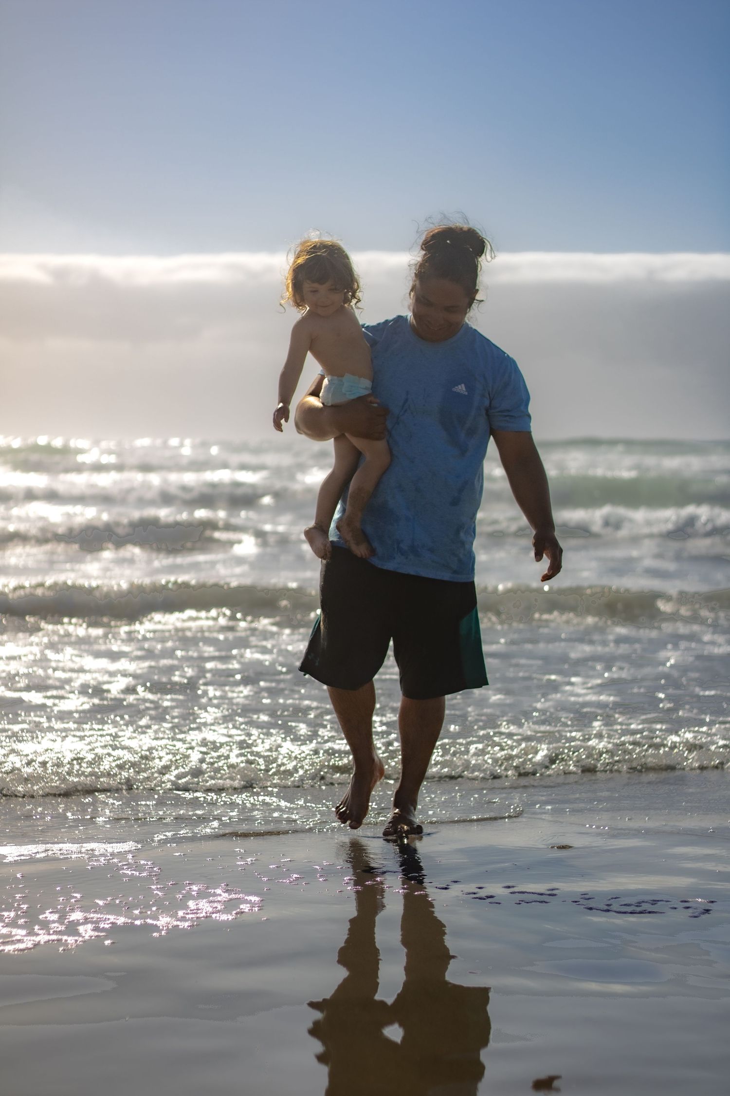 A man in a blue shirt carries a toddler while walking barefoot on a sunny beach with ocean waves behind them.