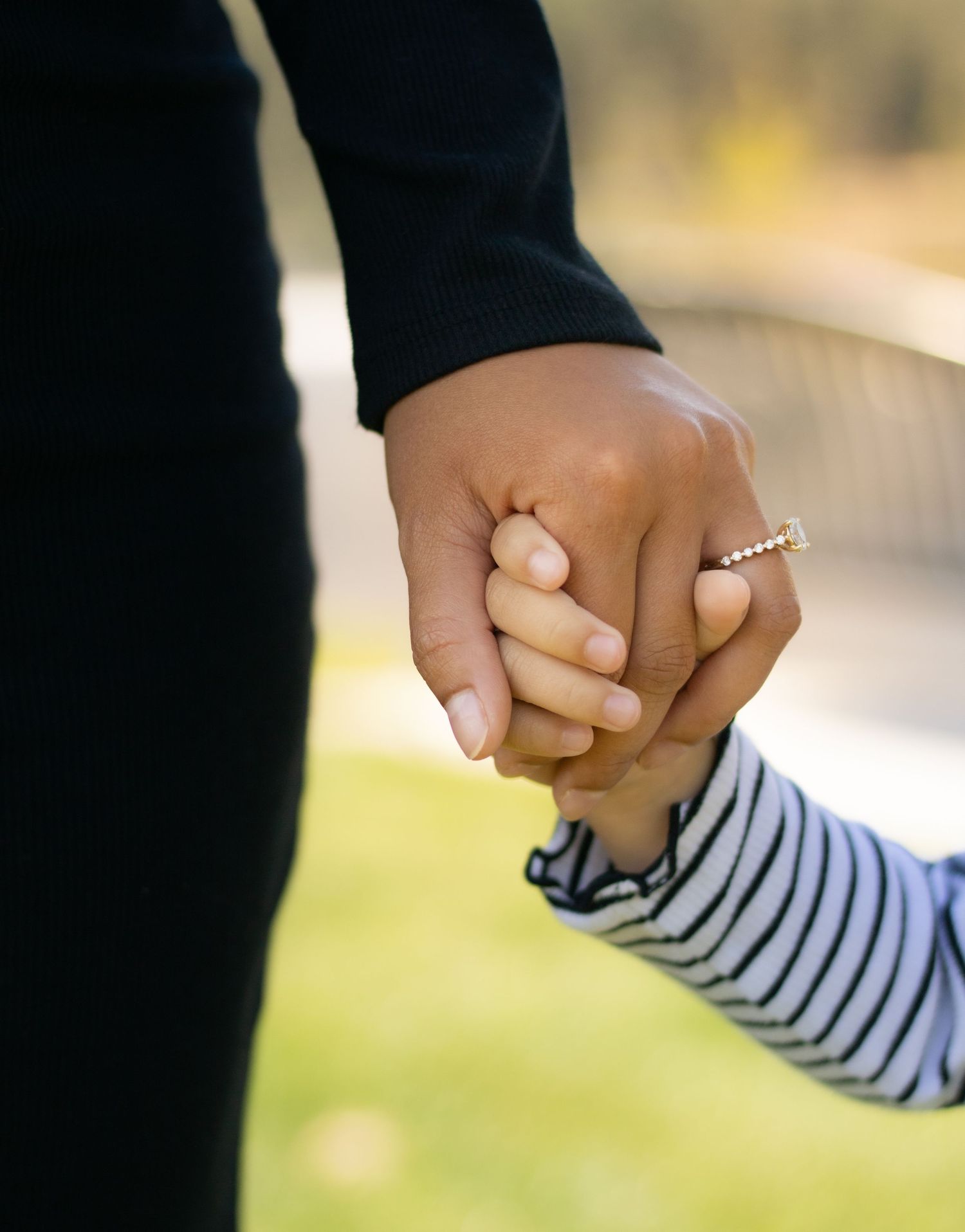 Adult holding a child's hand outdoors, wearing a diamond ring and black sleeve, with a blurred green background.