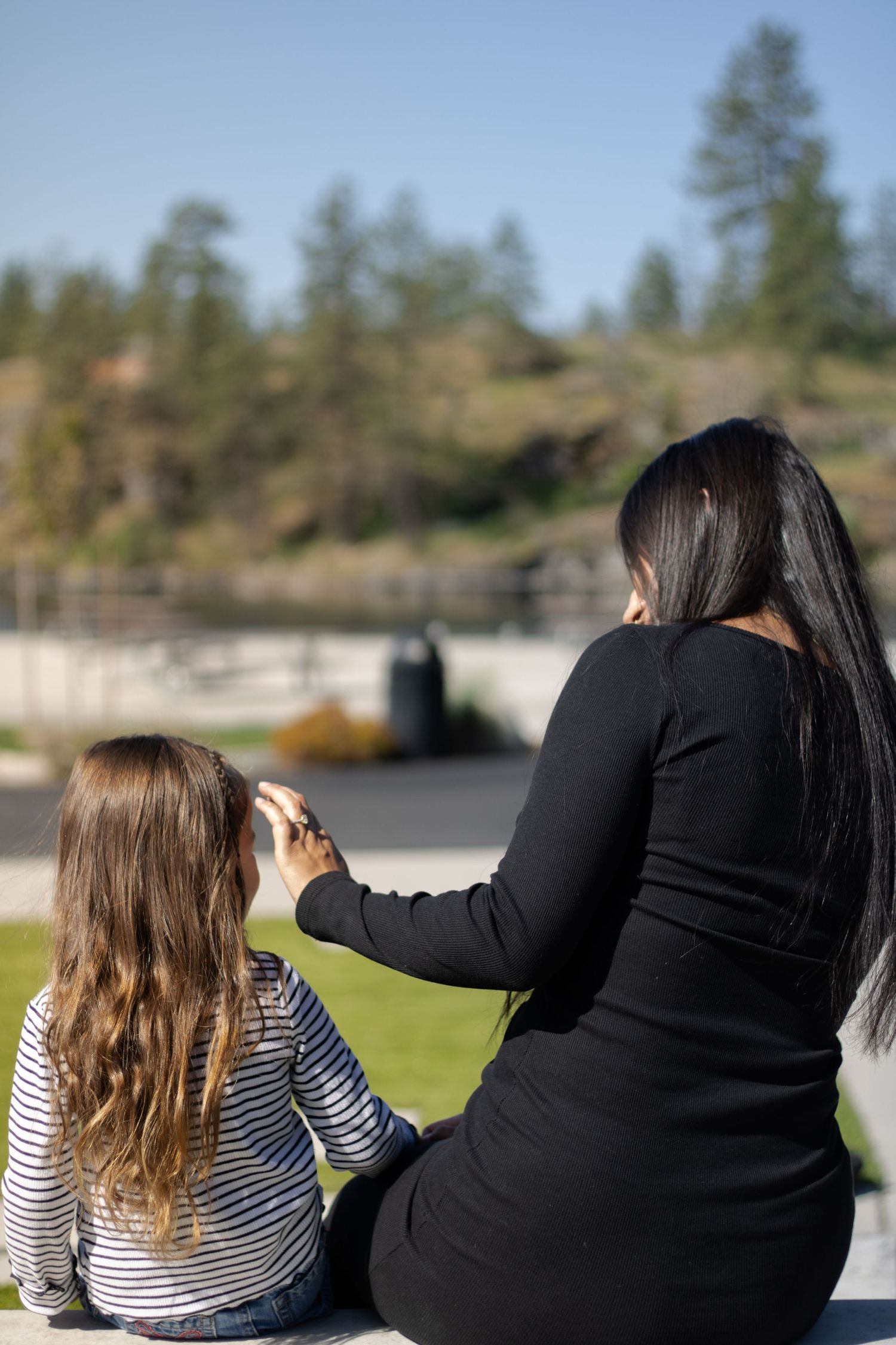 A woman in black dress sits with a young girl, pointing at a scenic park view with pine trees and blue sky.