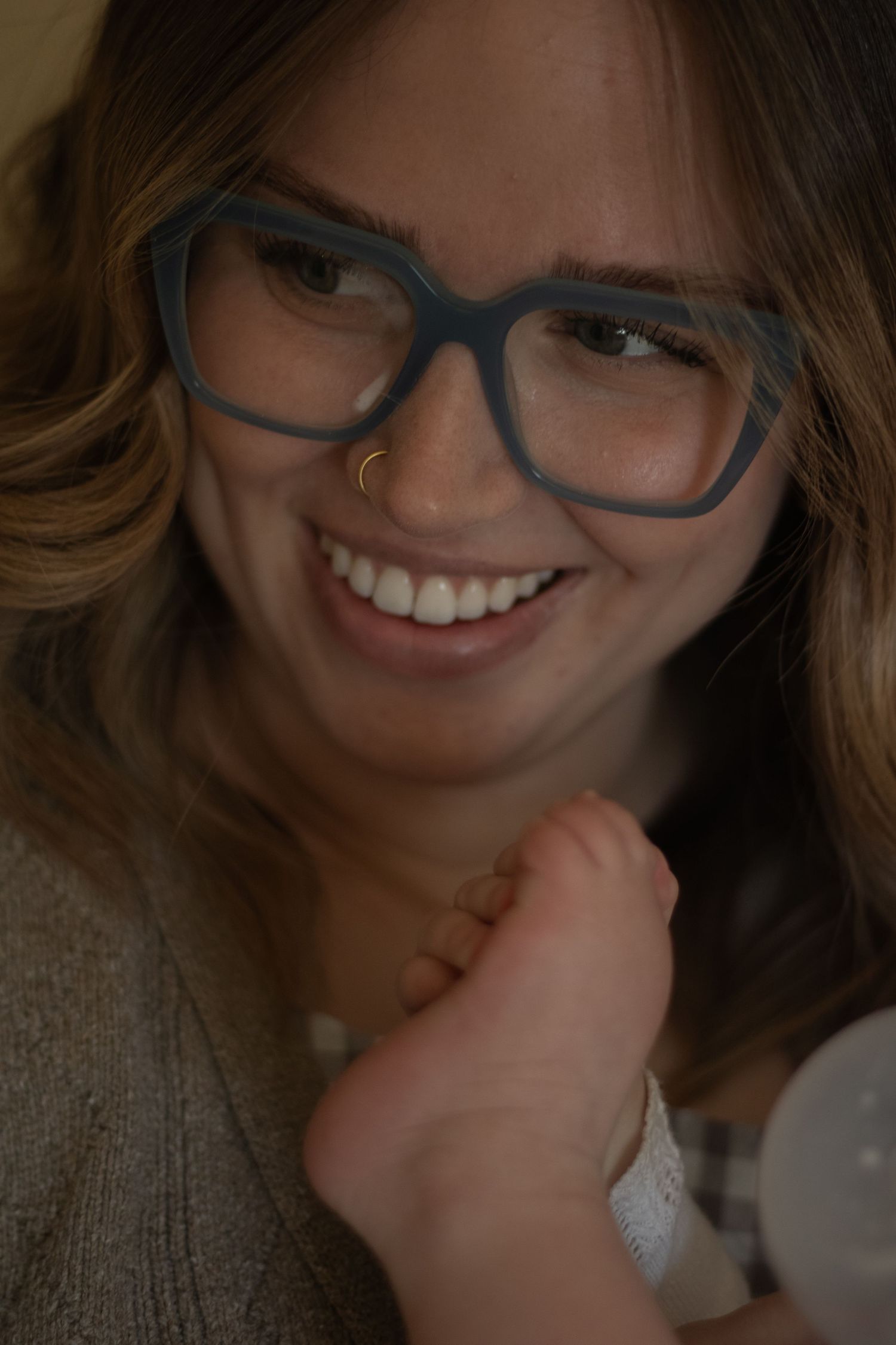 Smiling woman with blue geometric glasses and nose ring holds a baby's foot close to her face.