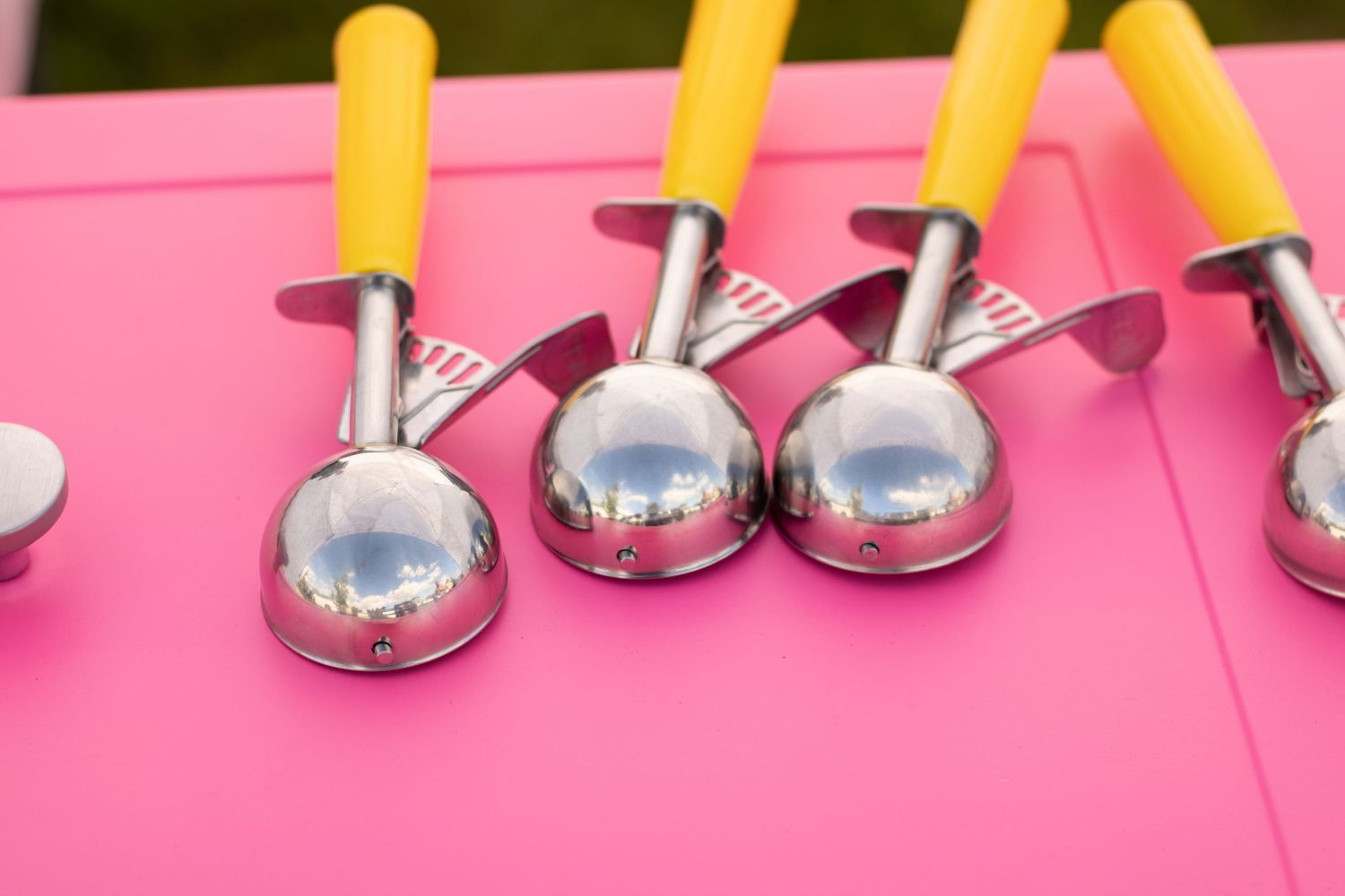 Shiny stainless steel ice cream scoops with yellow handles resting on a vibrant pink surface.