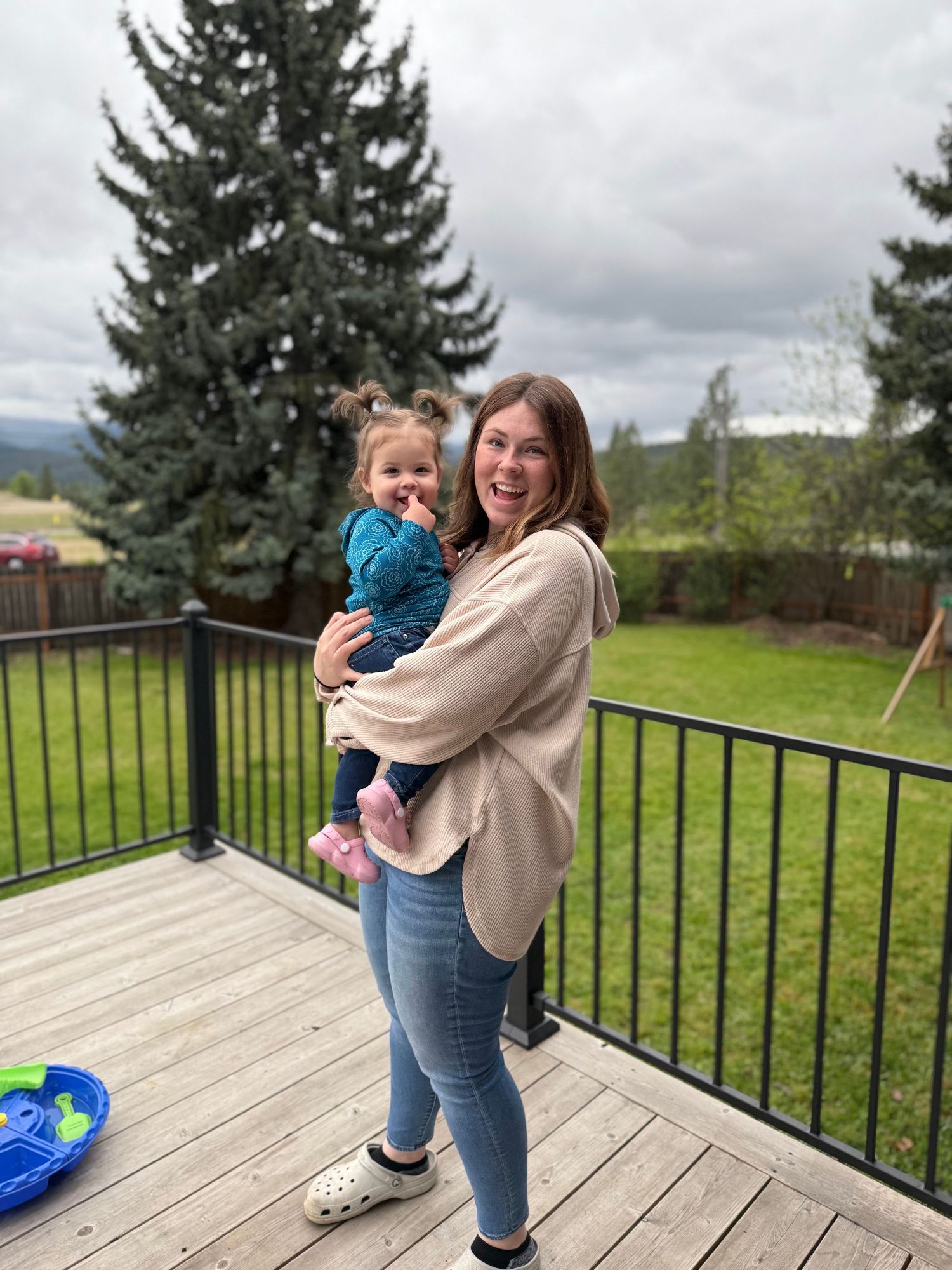 A smiling woman holds a toddler girl on a wooden deck with a lush green lawn and tall evergreen tree behind them.
