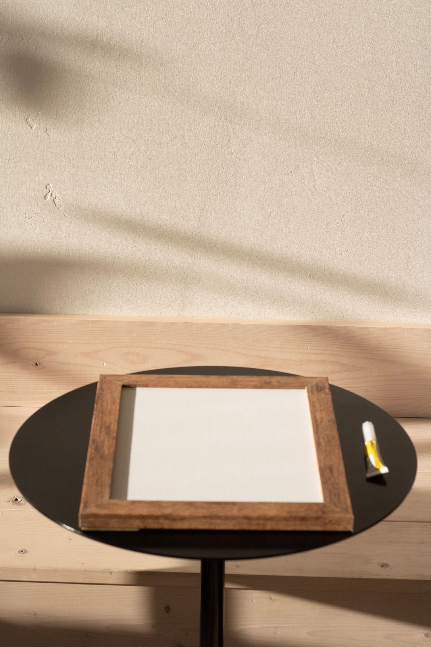 Wooden frame with blank white canvas on black round table, yellow marker beside it, natural light shadows on beige wall.