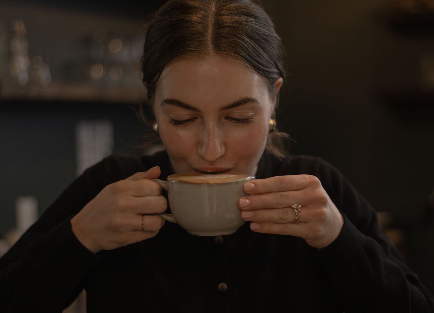 Young woman with closed eyes sipping coffee from a ceramic cup in a cozy cafe setting.