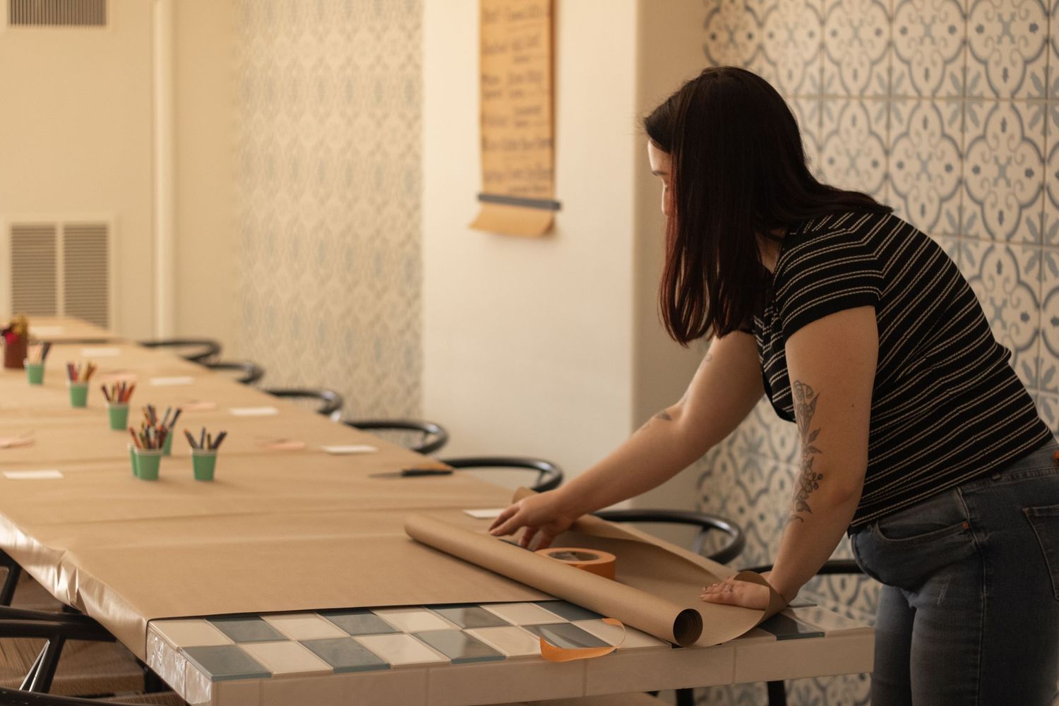 Woman rolling out paper on a long craft table in an art studio with colorful pencils and tiled walls.