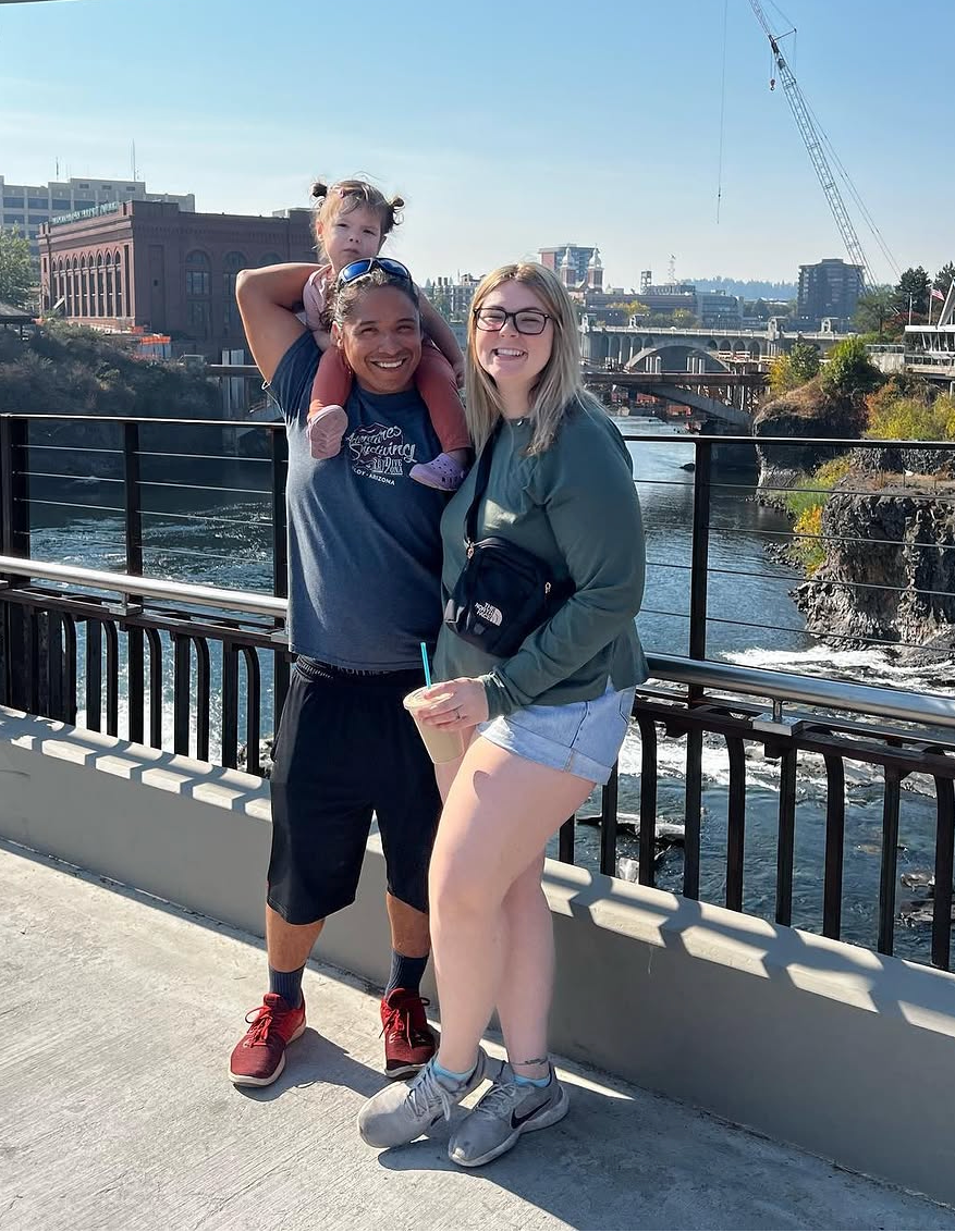 A couple smiles on a bridge over a river, with a toddler on shoulders, in a sunny urban setting.