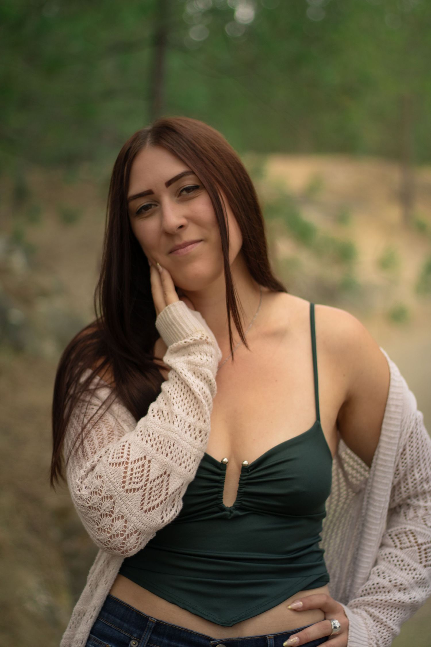 Woman with long brown hair in green top and white knit cardigan posing outdoors in forest setting.