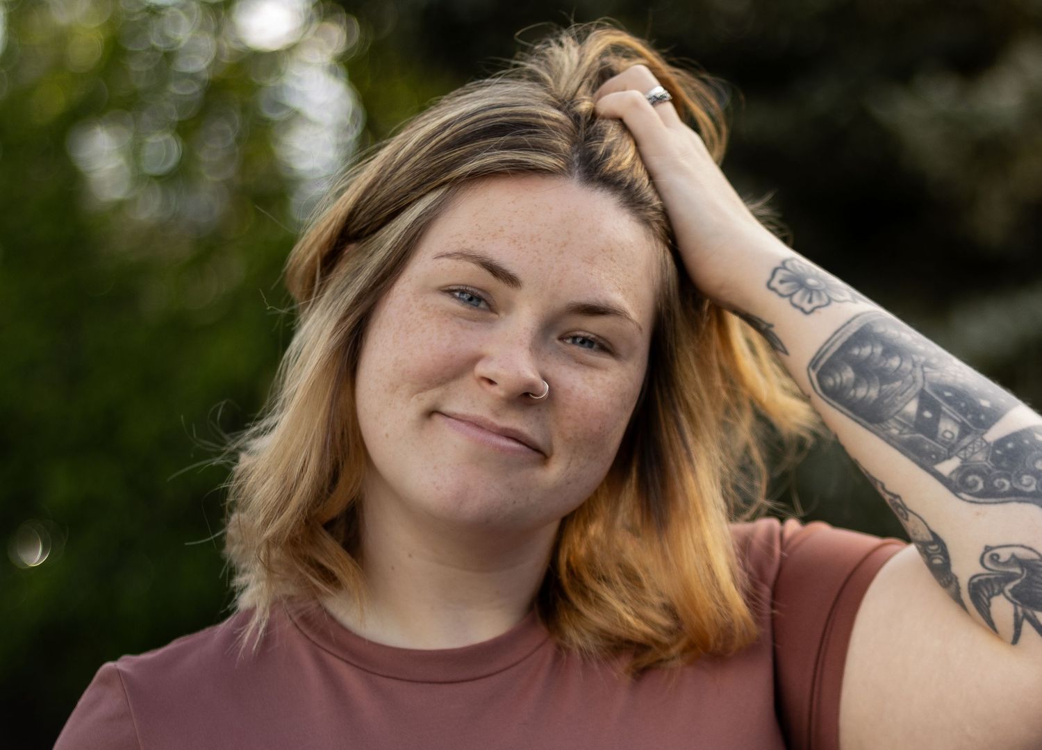 Young woman with freckles and arm tattoo smiling outdoors, hand in blonde hair, wearing a mauve t-shirt.