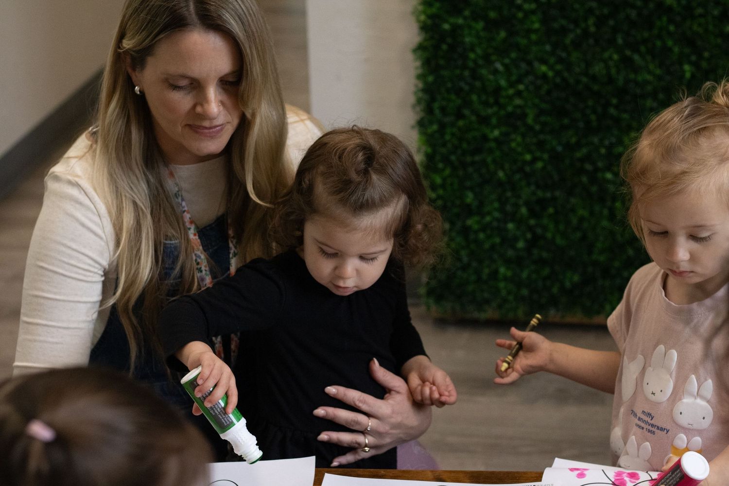 Woman helps toddler with craft activity at table while another child participates in kids art project.