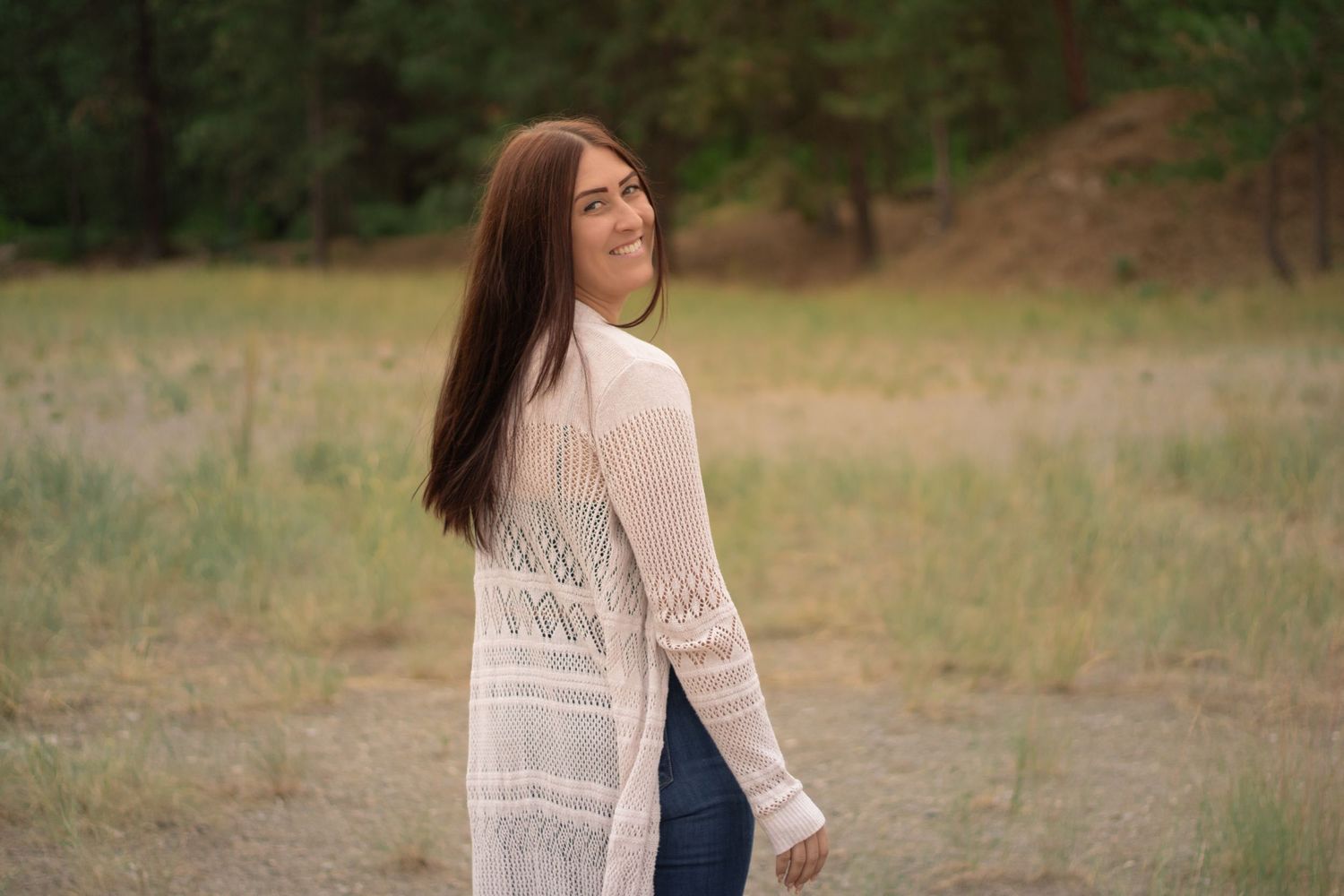 Young woman with long brown hair in white cardigan and jeans smiling outdoors in a natural field setting.
