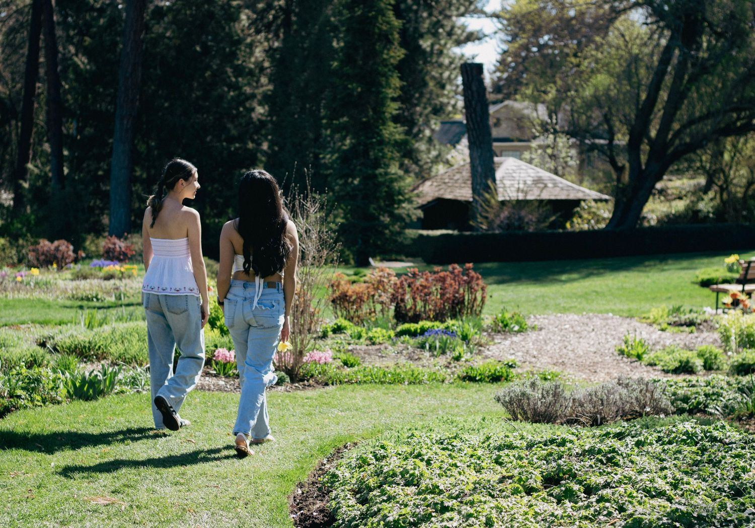 Two women walking hand in hand through a lush garden with colorful flowers and trees on a sunny day.