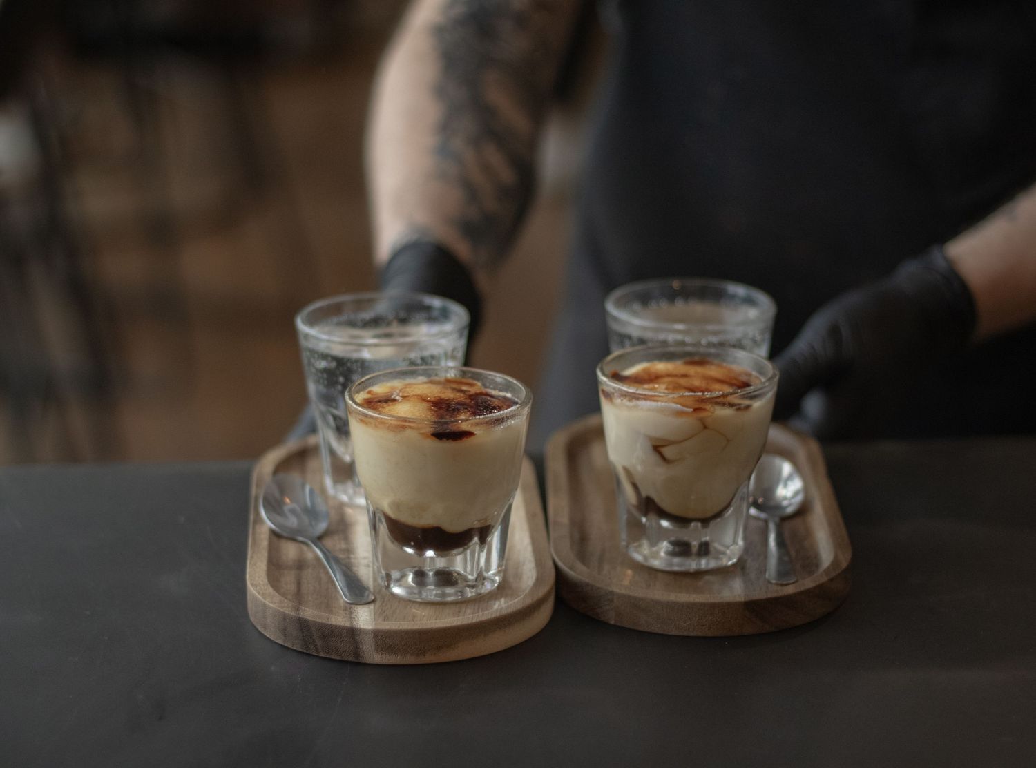 Tattooed barista serving two espresso tiramisu dessert drinks on wooden trays in glass cups.