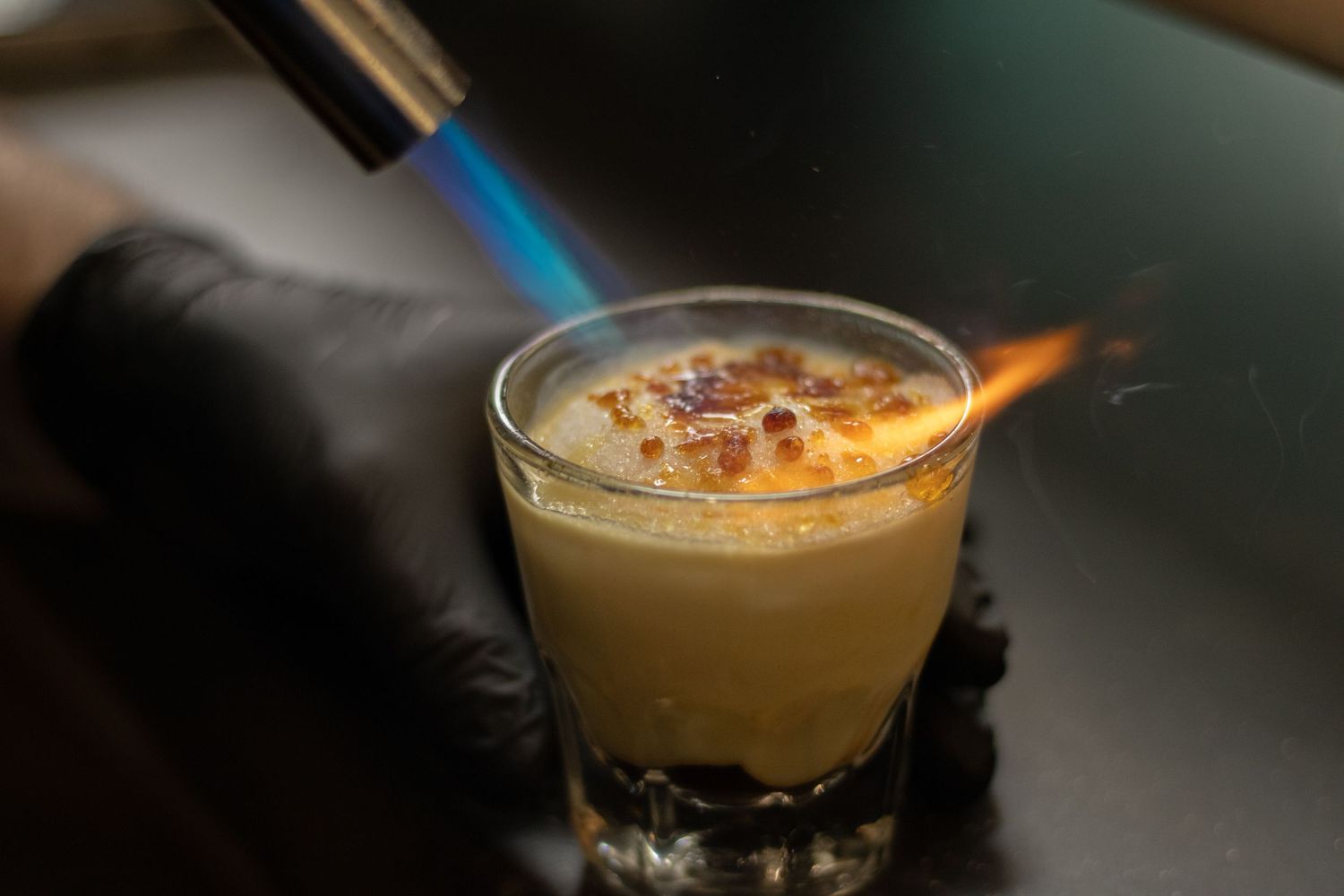 Bartender torching a creamy cocktail shot with a blue flame torch while wearing black gloves.