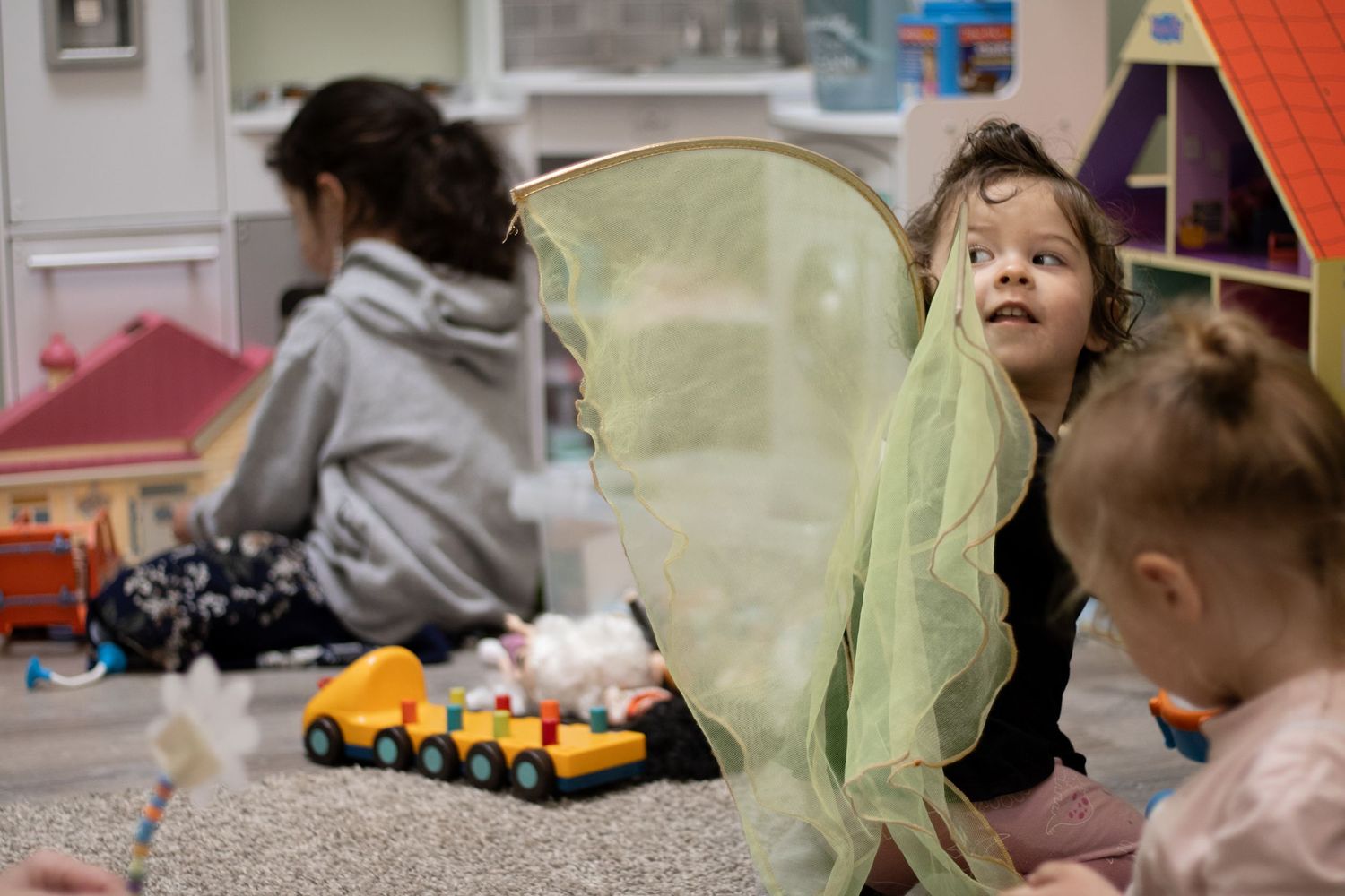 Children playing in a colorful daycare classroom, with a toddler holding a green fairy wing while sitting on the floor.