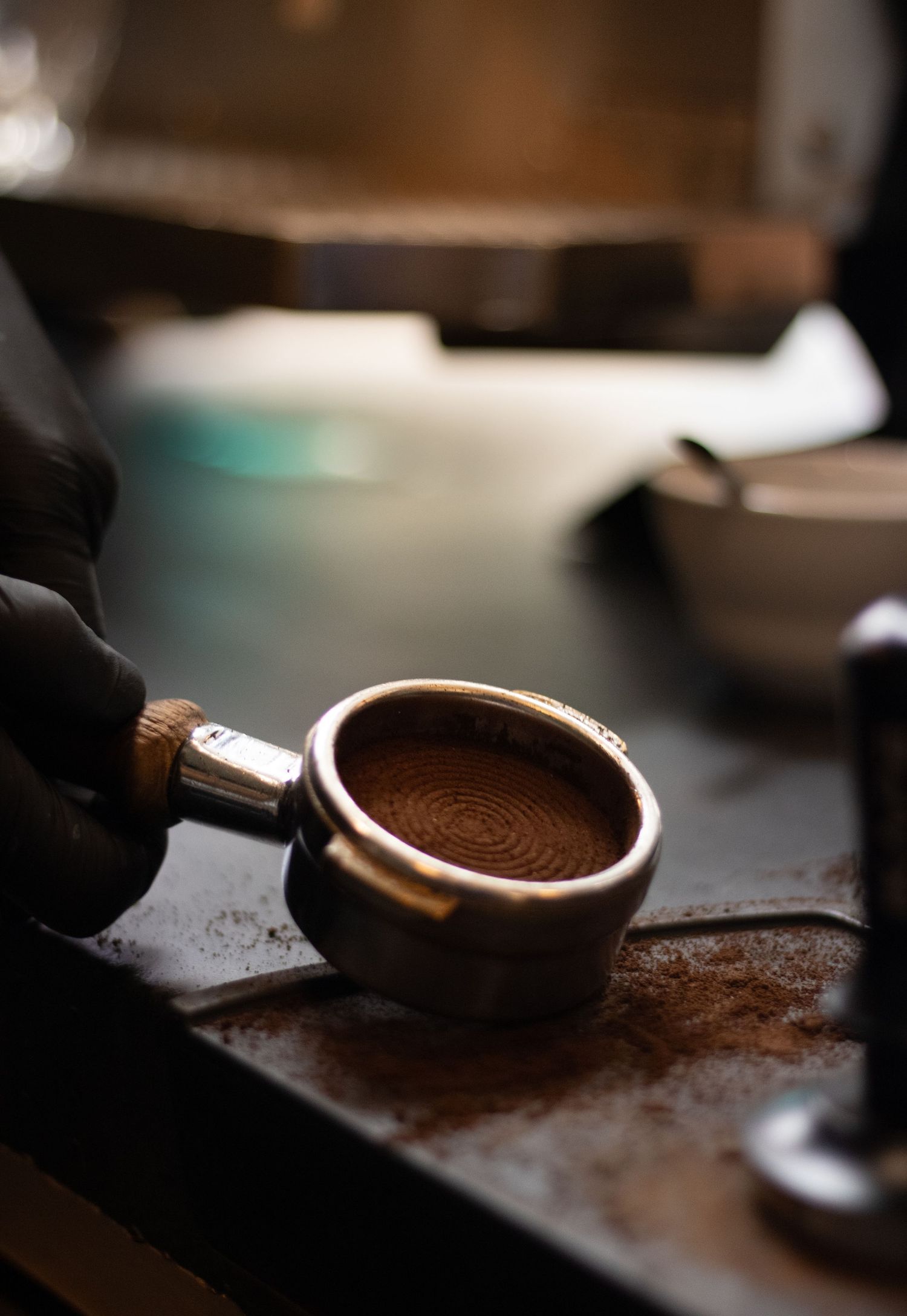 Barista in black gloves holding a portafilter filled with freshly tamped espresso grounds on a coffee bar.