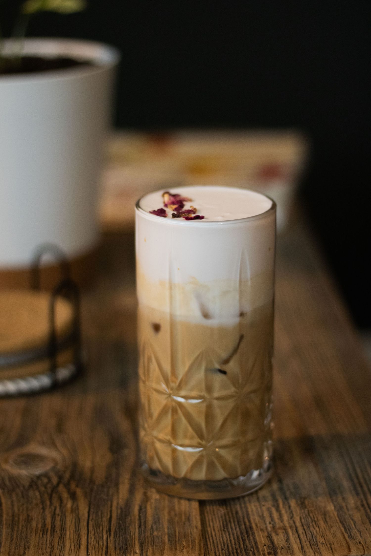 Iced latte in a tall geometric glass topped with foam and dried rose petals on a rustic wooden table.