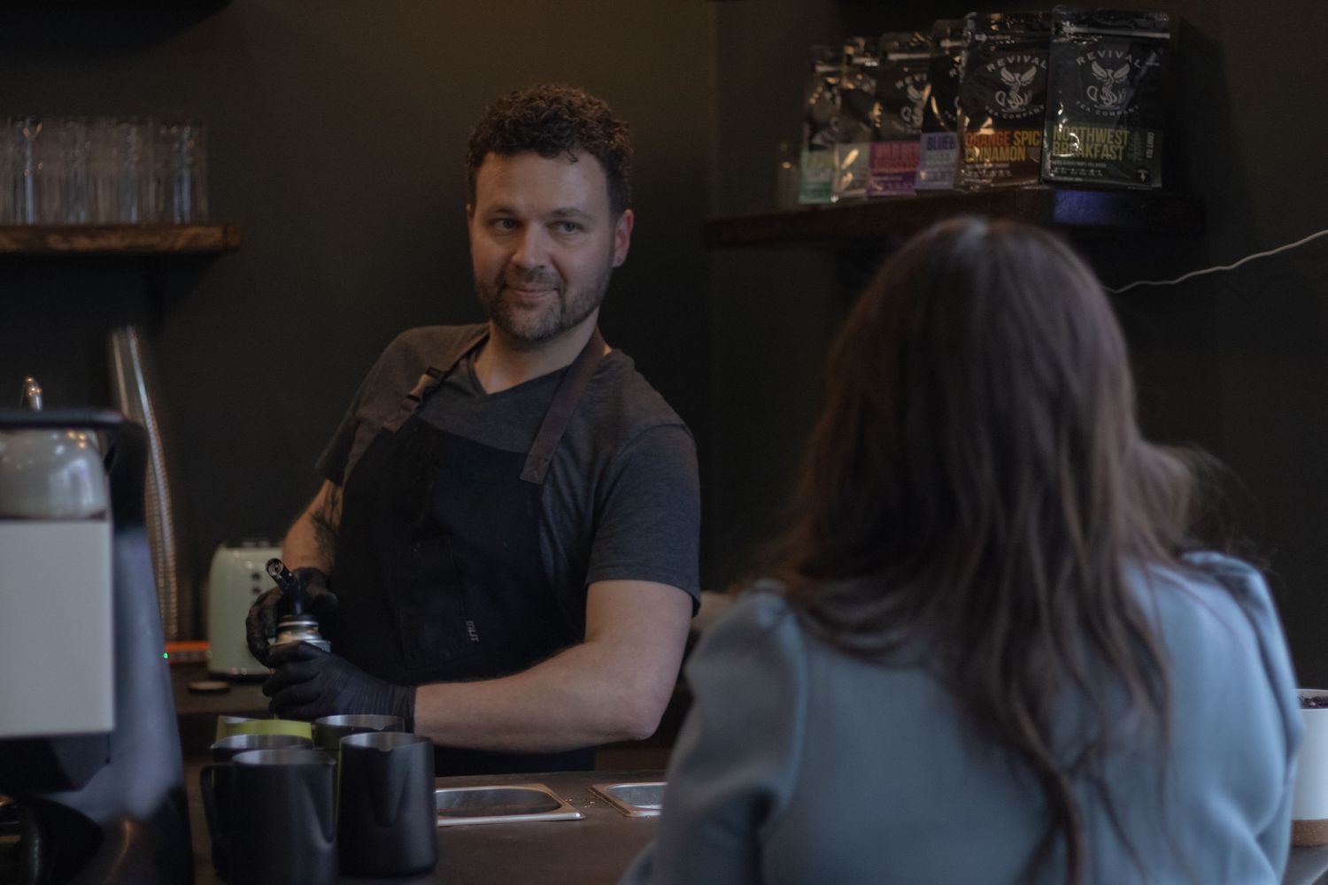 Barista in black apron smiling at female customer across coffee shop counter in dimly lit cafe.