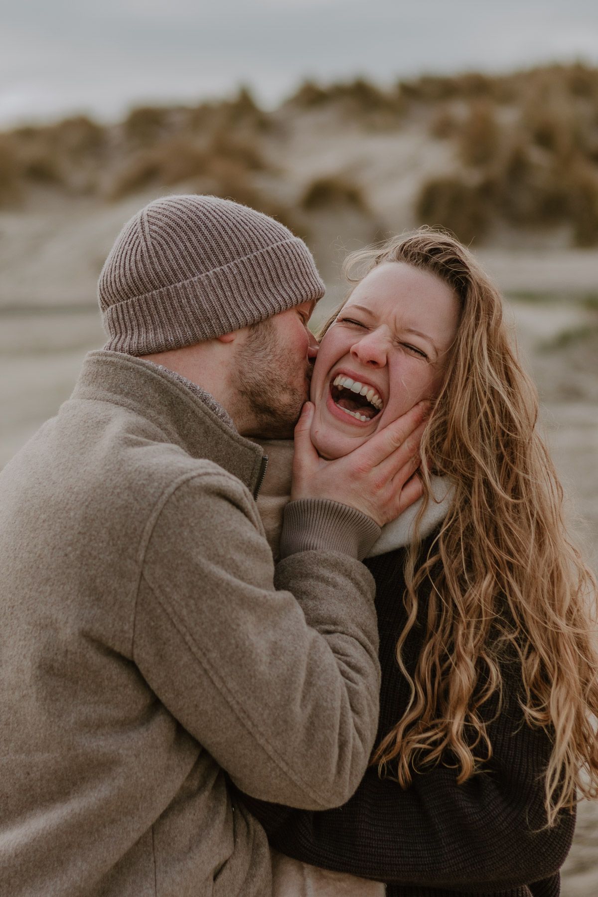 A joyful couple sharing a tender moment on a windy beach, the woman laughing as her partner kisses her cheek.