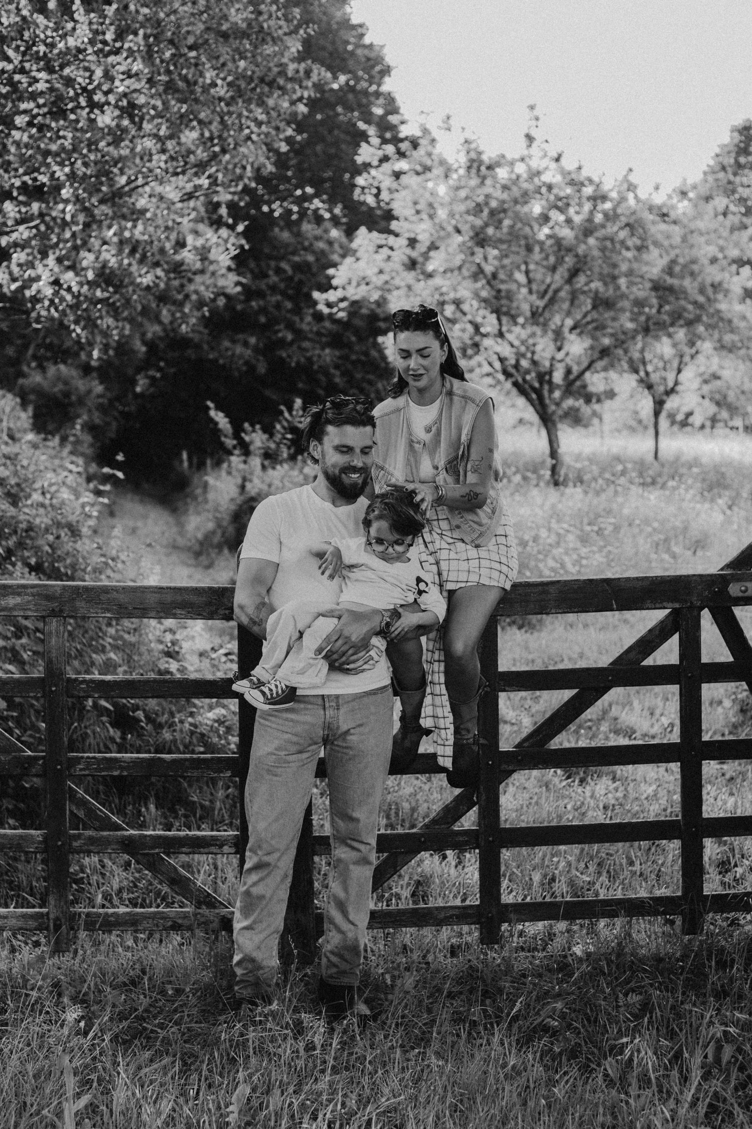 Black and white family portrait of couple with young child by rustic wooden gate in countryside.