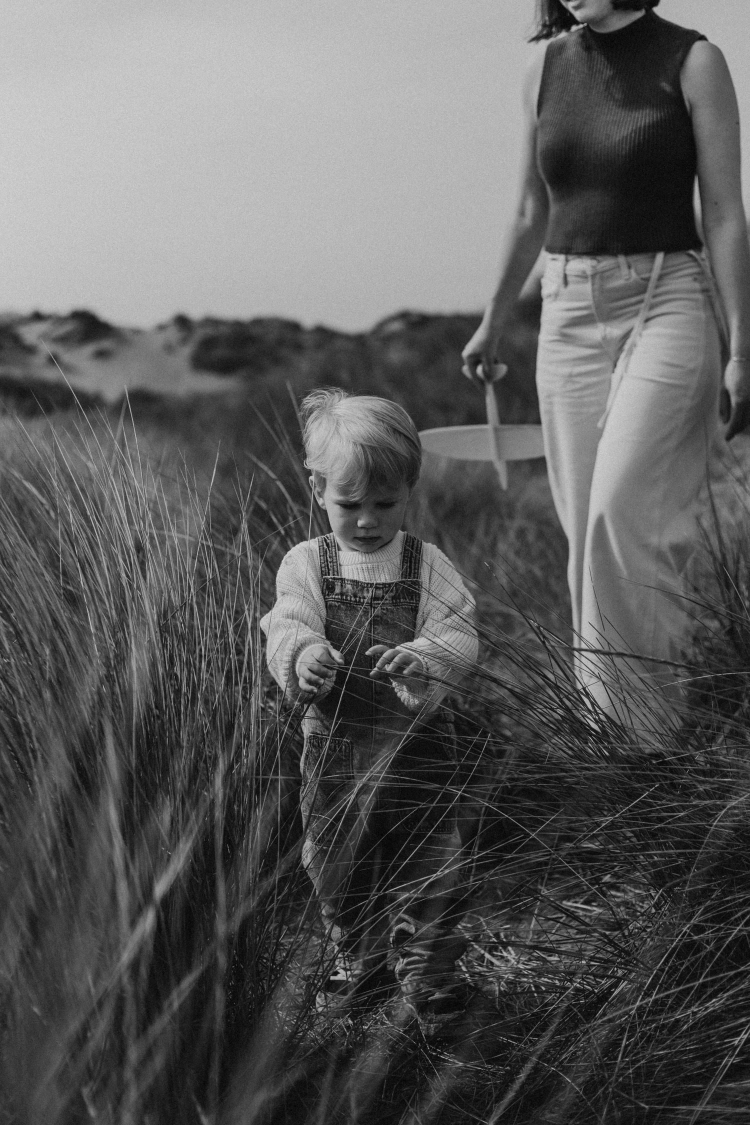 Black and white photo of toddler exploring tall grass while mother walks nearby.