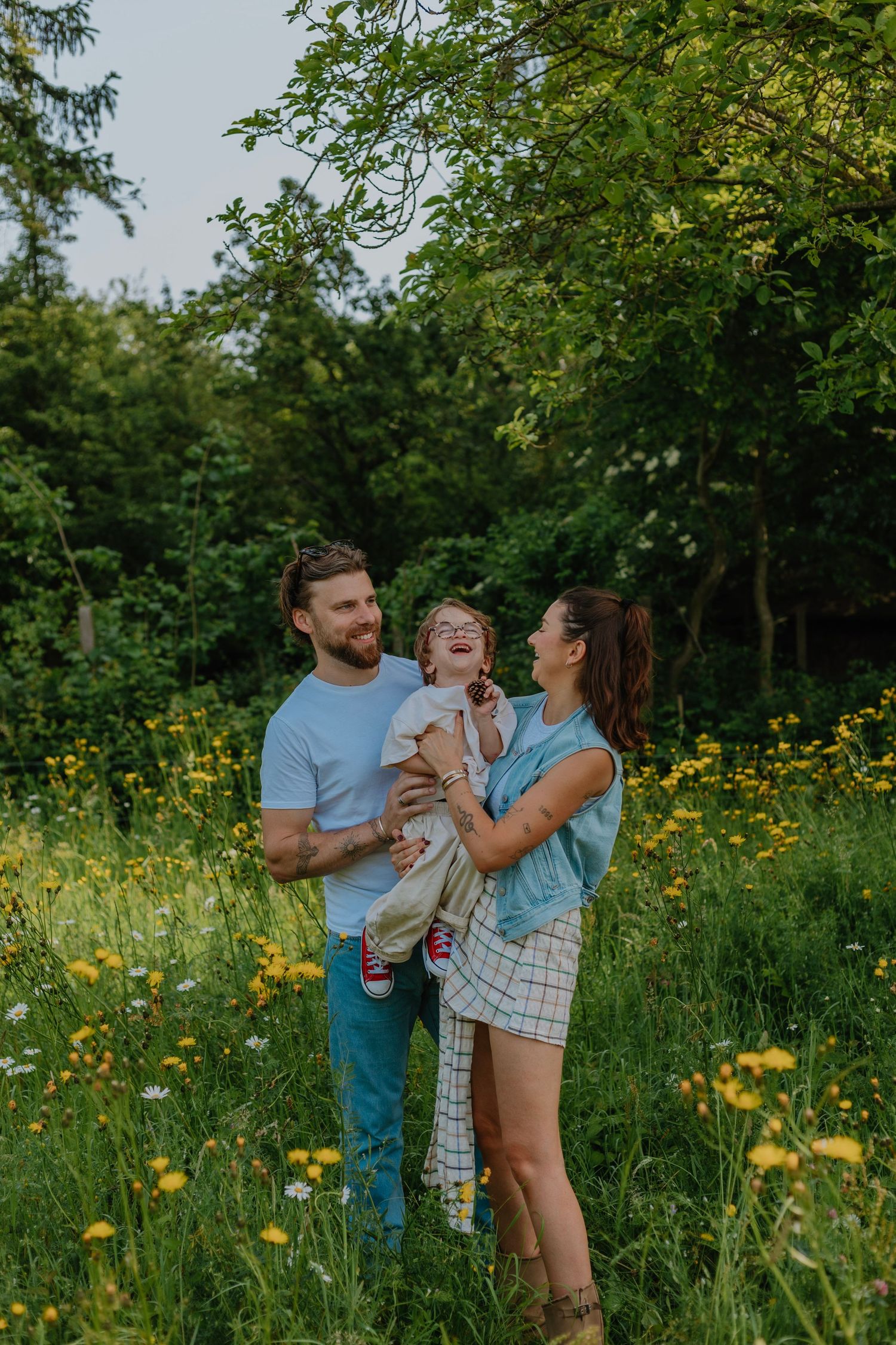 Happy young family with a laughing child standing in a sunny wildflower meadow surrounded by lush green trees.