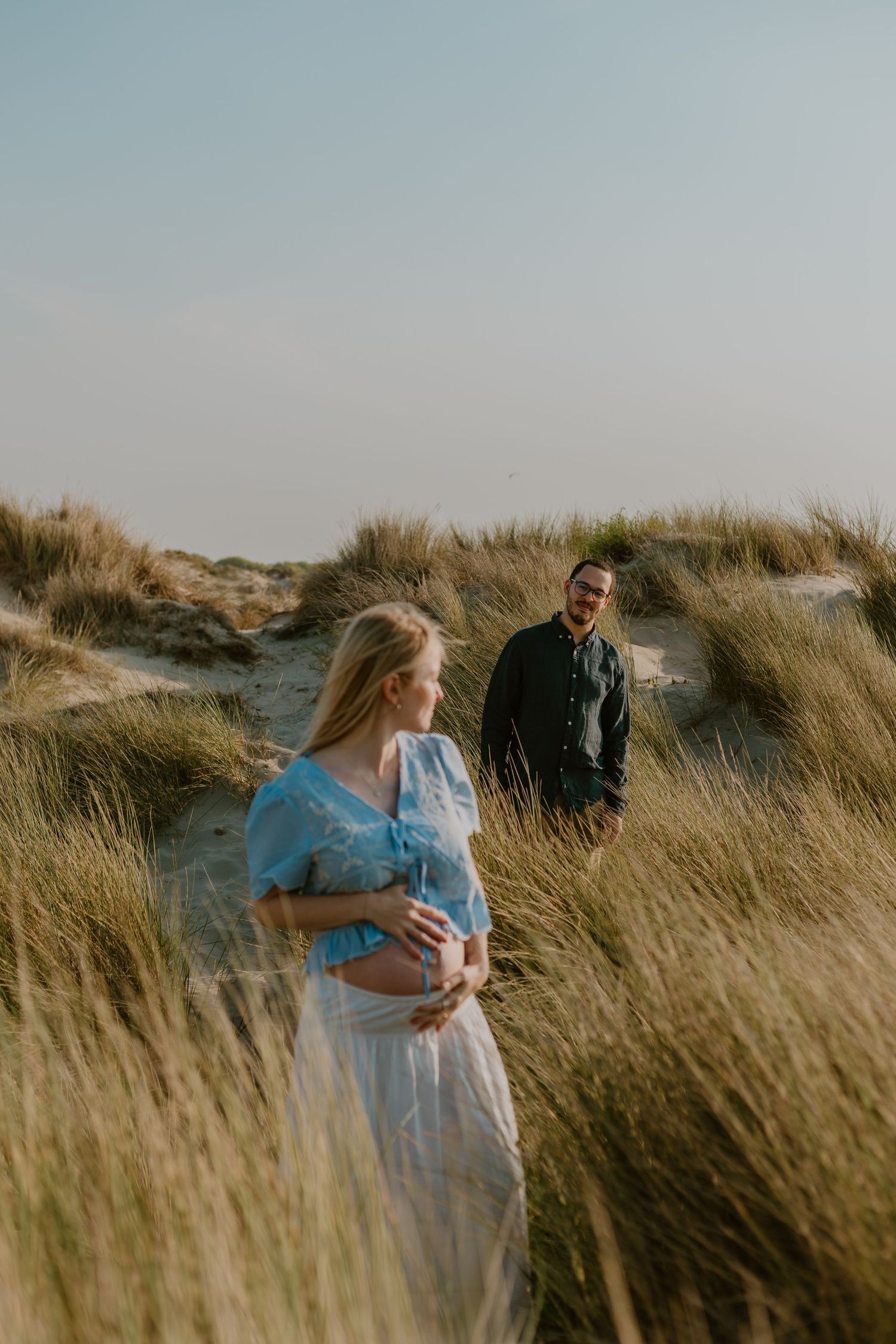 Pregnant woman in blue dress holds belly in coastal dunes as man smiles at her from behind tall grass.