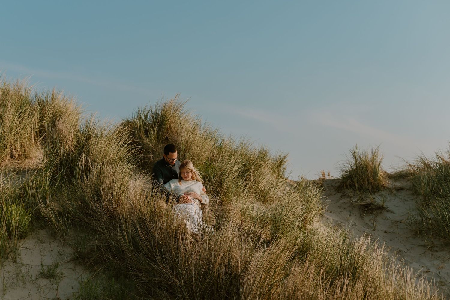 Romantic couple embracing among tall grass on sun-kissed sand dunes under blue sky.