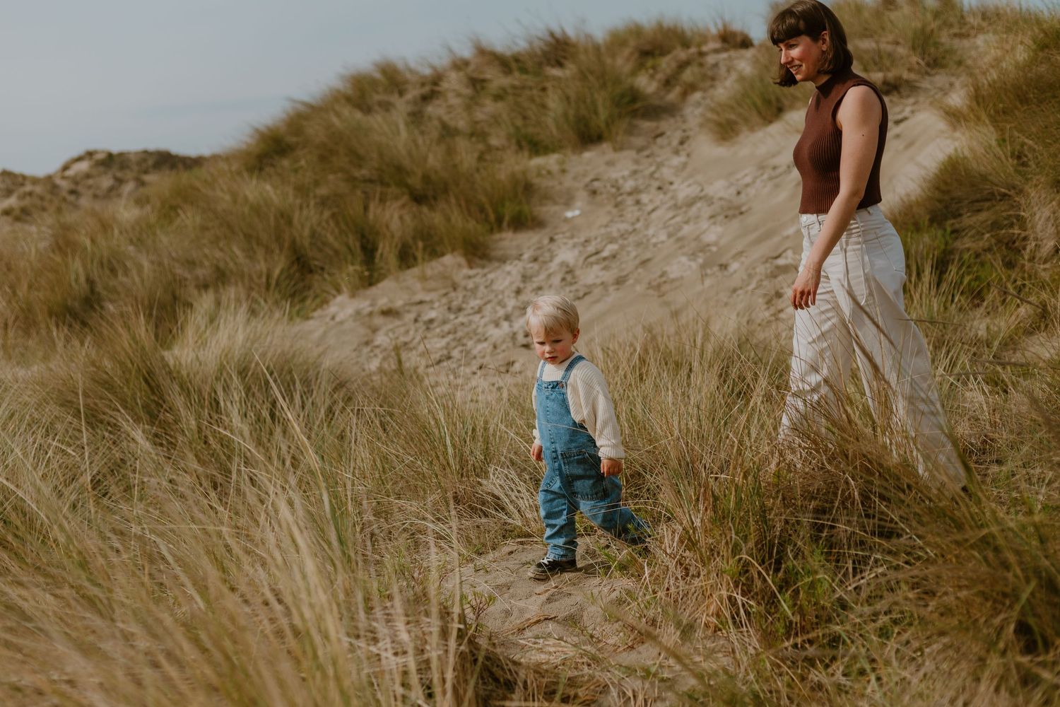 Mother and toddler walking through sandy beach dunes with tall grass.