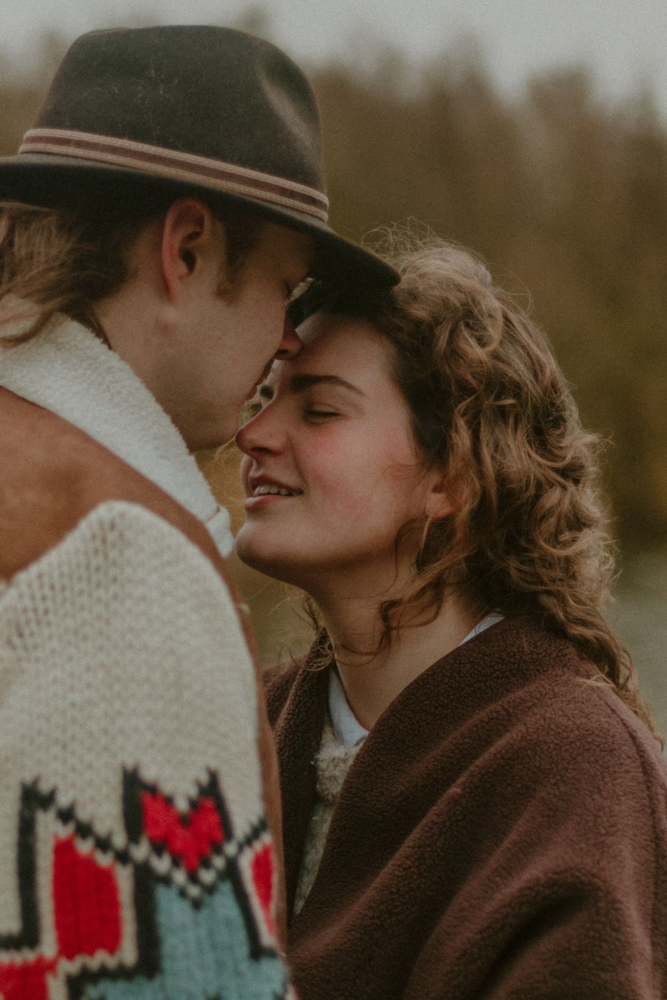 Close-up of a smiling woman leaning toward a man in a patterned sweater and hat outdoors.