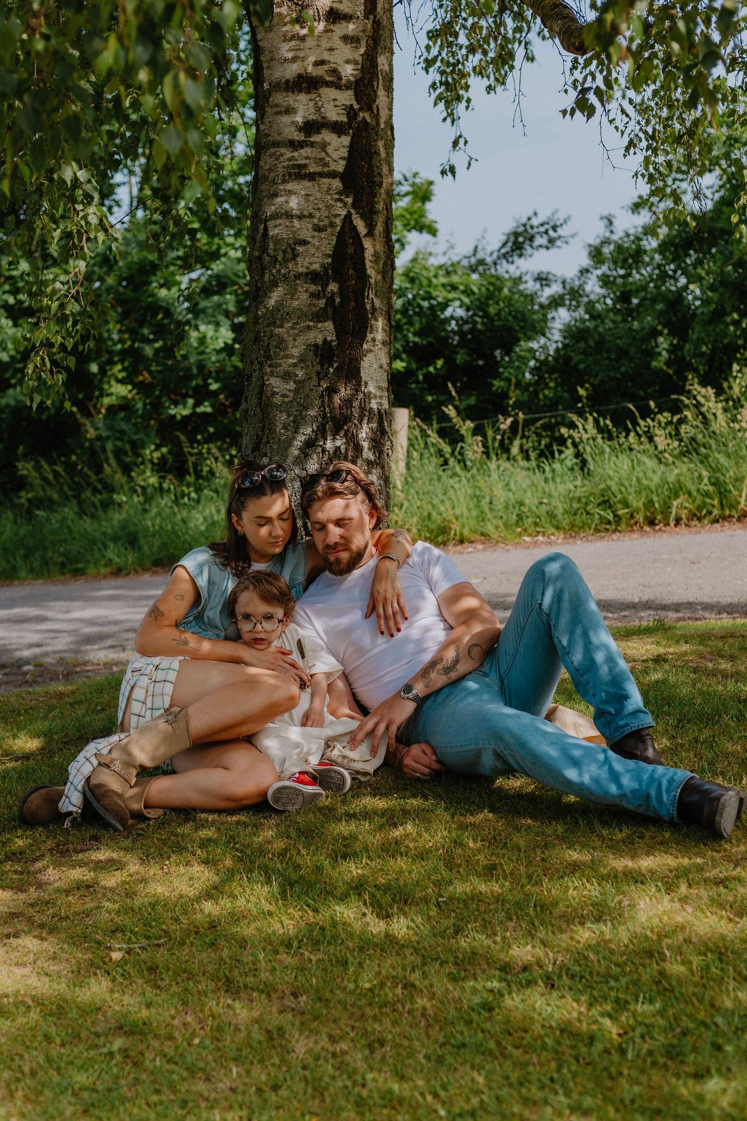 Family of three relaxing under a tree in a park, parents reading with young child on sunny day.