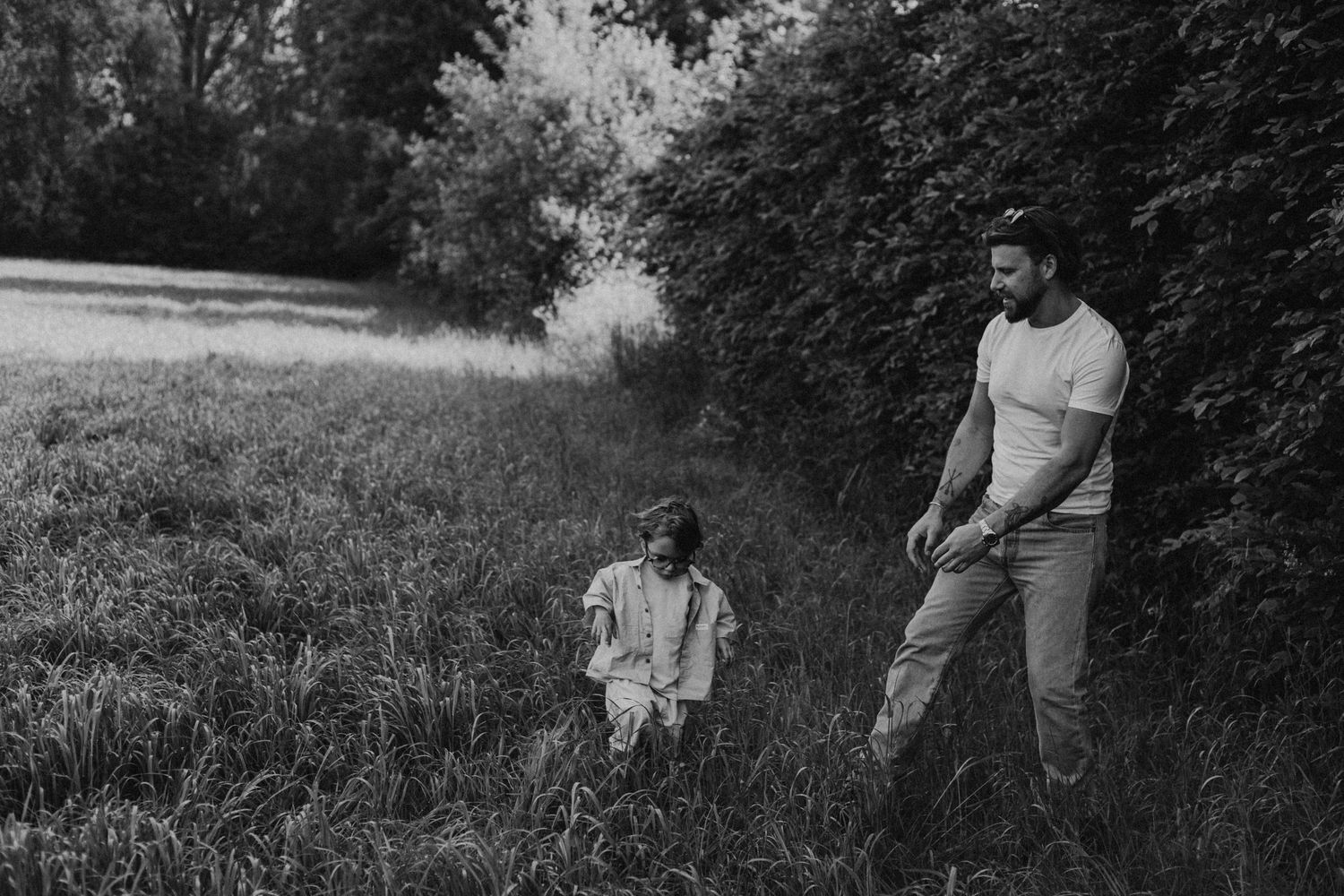 Black and white photo of a father and young child playing together in a lush green meadow near trees.