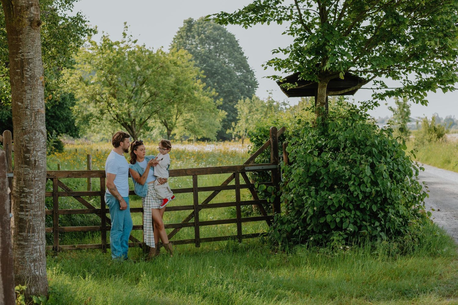 Family of three standing by a rustic wooden gate surrounded by lush green trees in a countryside setting.