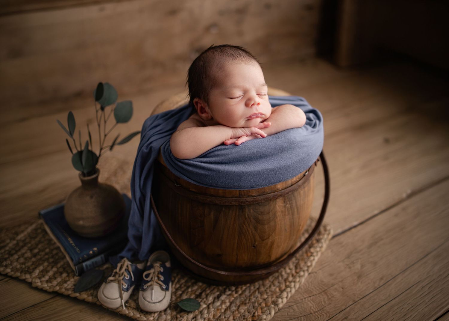Newborn baby sleeping peacefully in a wooden barrel wrapped in blue fabric, with tiny shoes and a vase nearby.