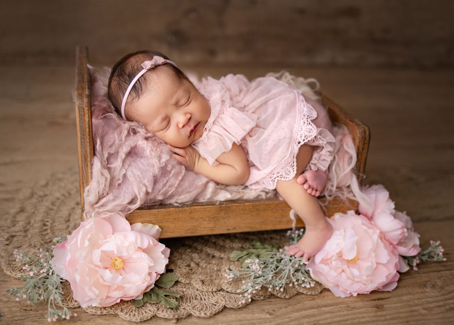 Newborn baby girl in pink lace outfit sleeping peacefully in a wooden mini bed with pink floral decorations.