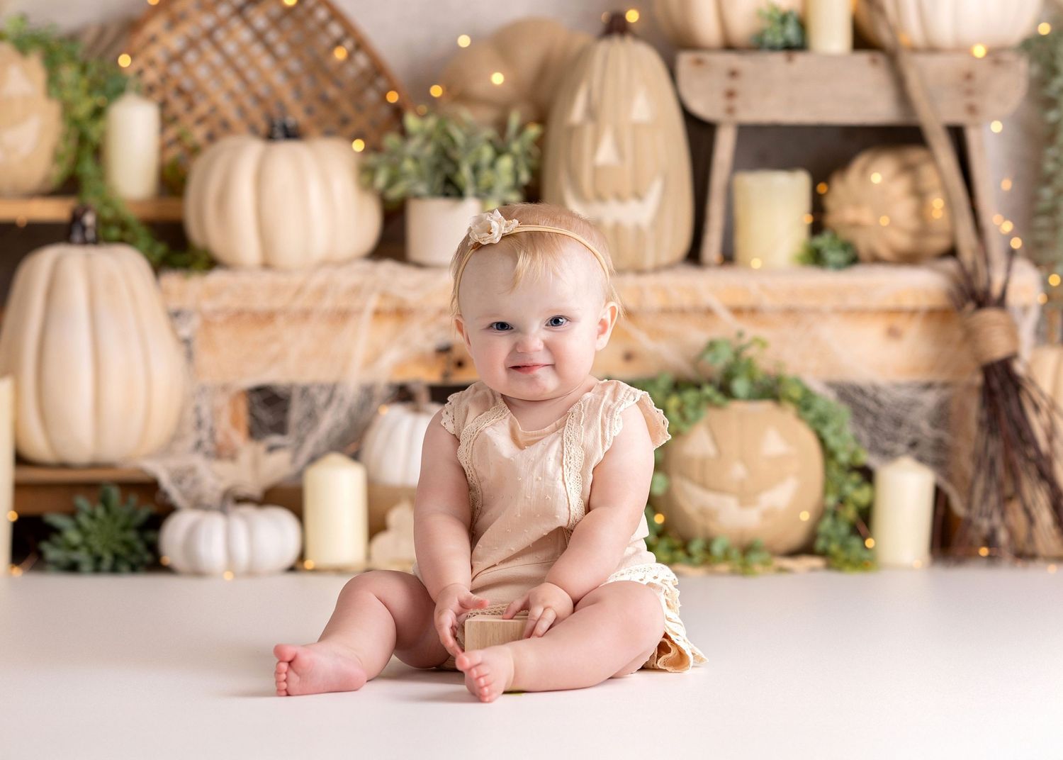 Smiling baby girl in beige romper sits before a fall backdrop with white pumpkins, neutral halloween, candles, and Halloween decorations.