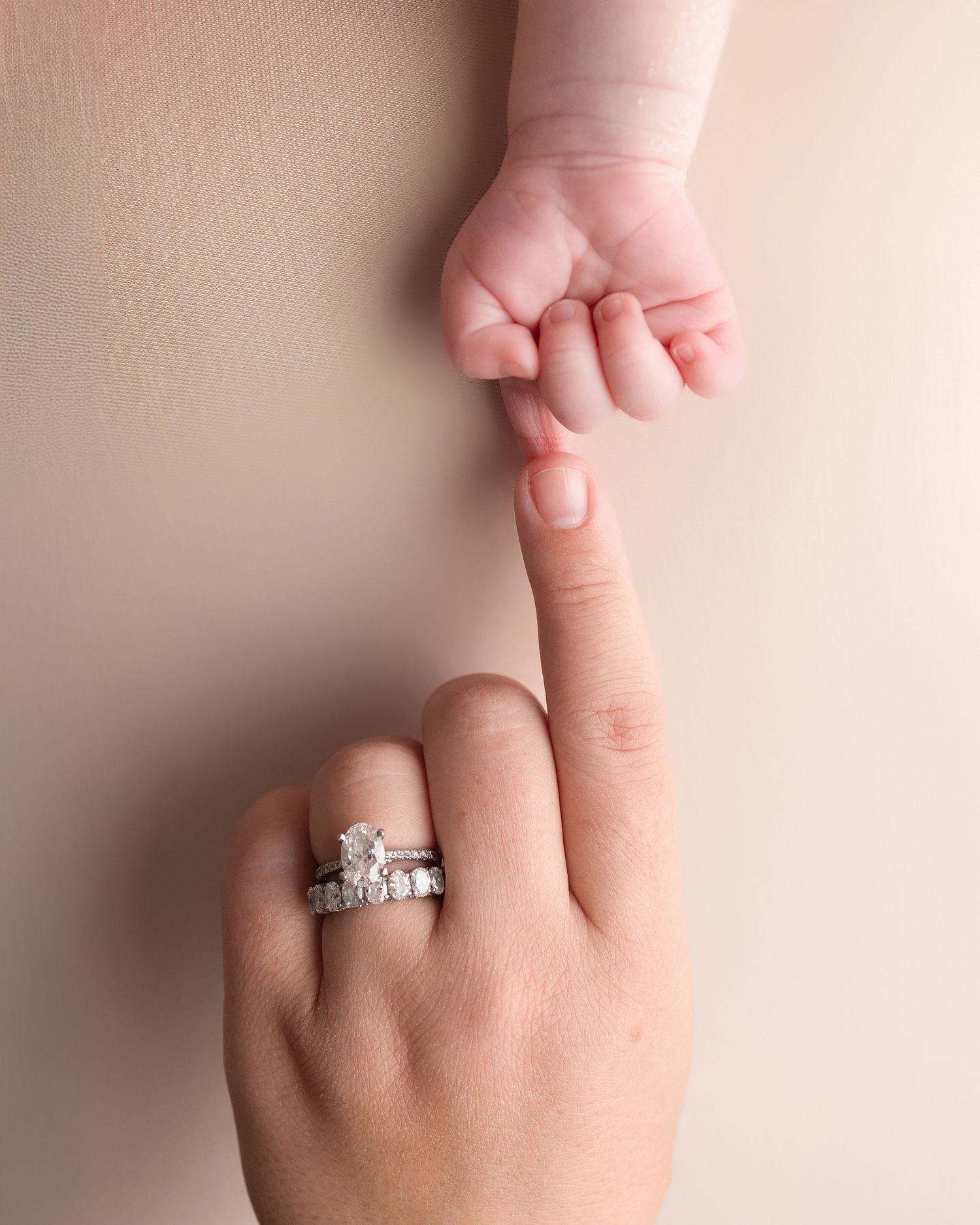 A newborn baby grasping an adult finger adorned with a diamond engagement ring and wedding band.