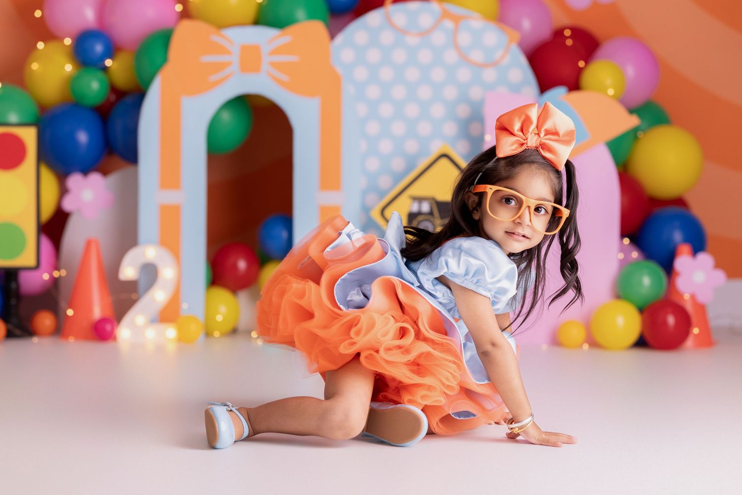 A young girl in an orange tutu and glasses poses playfully at a colorful balloon-decorated birthday party backdrop, blippi theme.