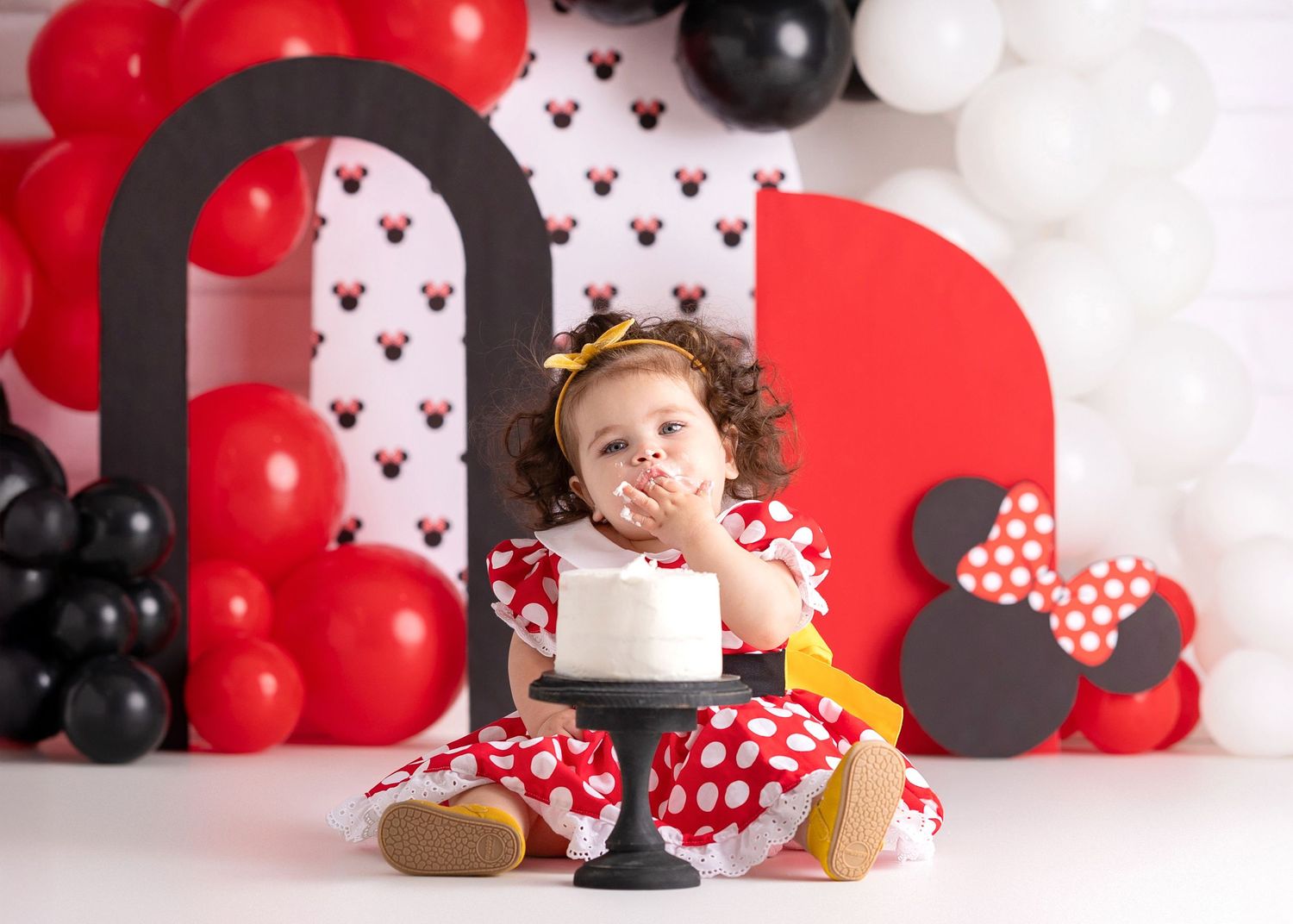 Toddler girl in red polka dot dress at Minnie Mouse themed birthday cake smash with balloons.
