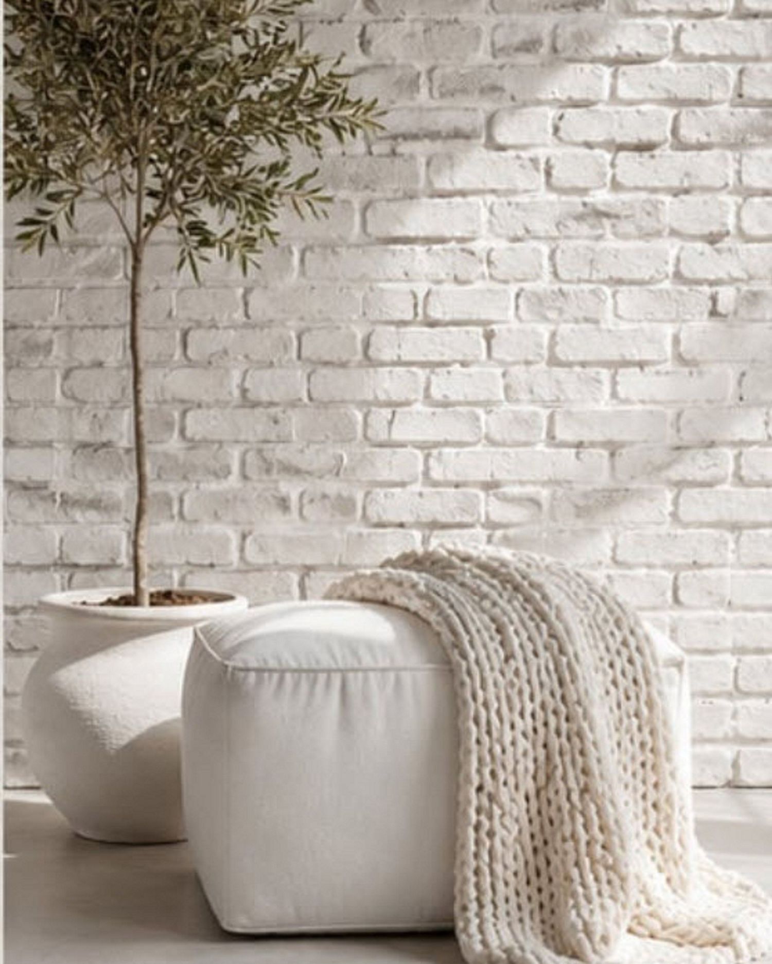 White brick wall interior with olive tree, round white pouf ottoman, and chunky knit throw blanket in minimalist decor.