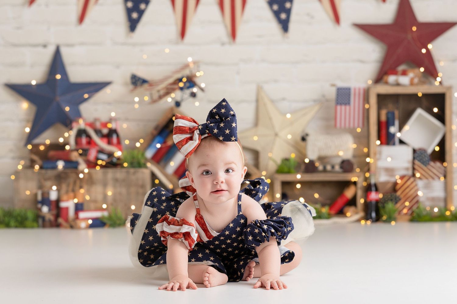 Baby girl in patriotic outfit crawls on white floor with American flag decorations and star backdrop, 250th anniversary.