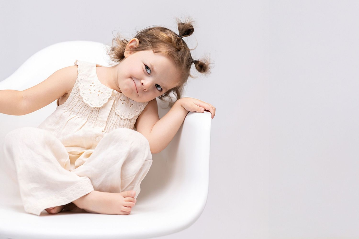 A cute little girl with pigtails in a white dress sits smiling in a modern white chair against a light background.