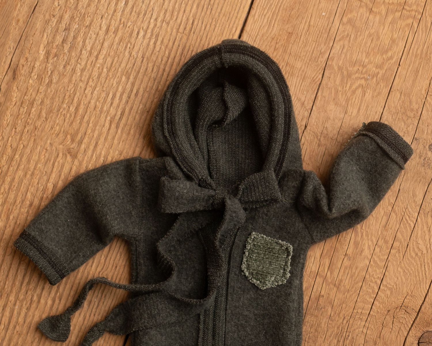 Dark olive green knitted baby hooded romper with tie strings and patch pocket, laid flat on wooden surface.