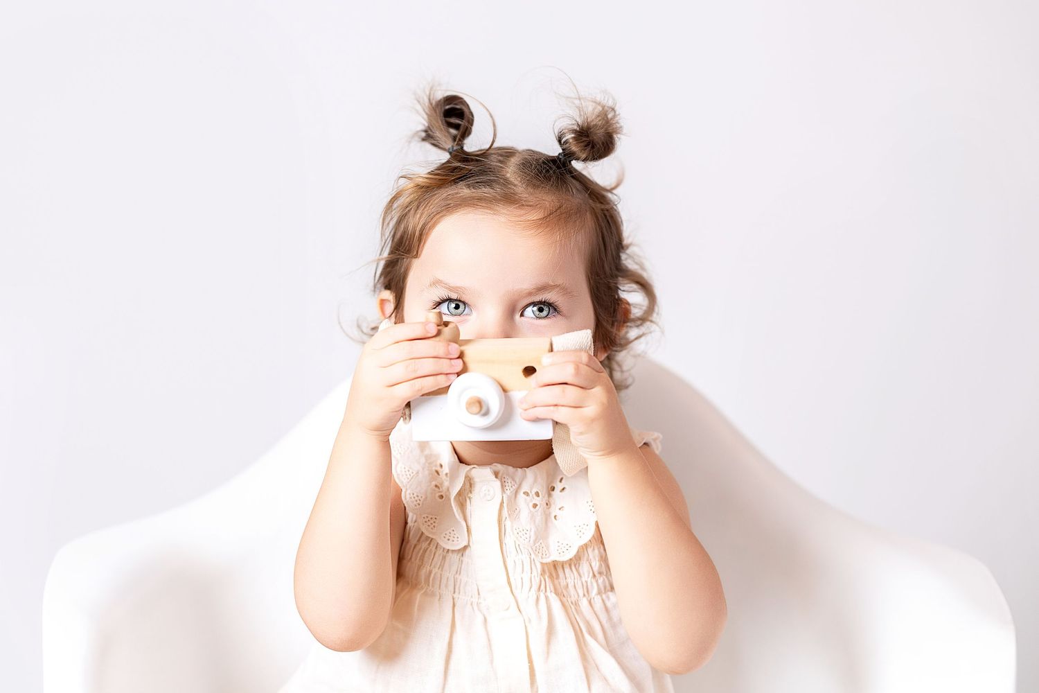 A cute toddler girl holds a small wooden toy camera up to her face while sitting in a white chair.